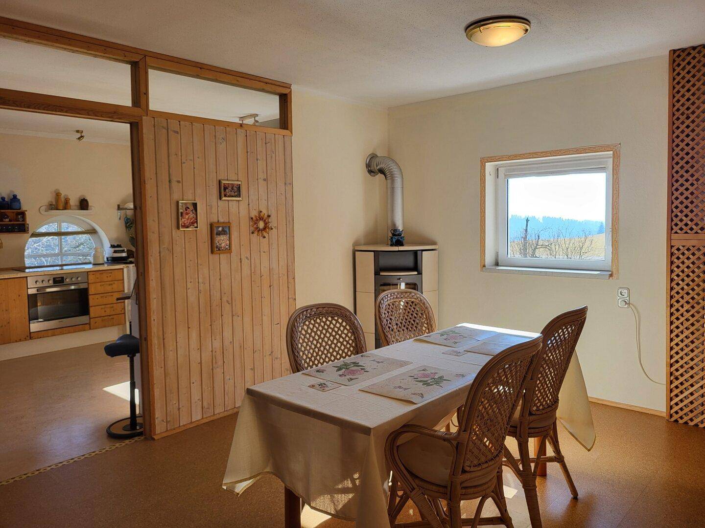 Essbereich mit Holztisch und Korbstühlen, neben einem Kachelofen und mit Blick in die Küche.
