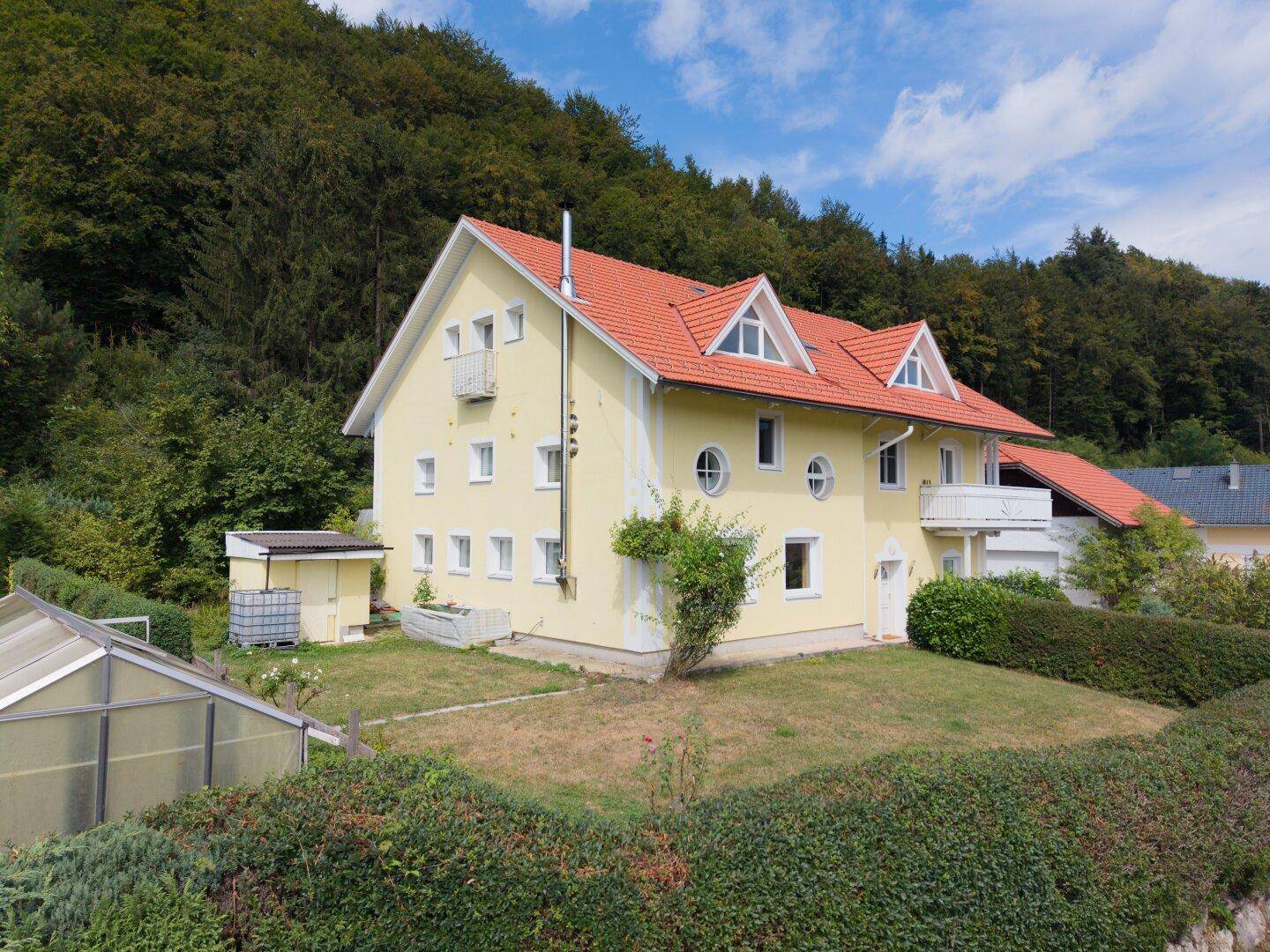 Luftaufnahme des Hauses von hinten mit Gewächshaus, Schuppen und umgebendem Wald.