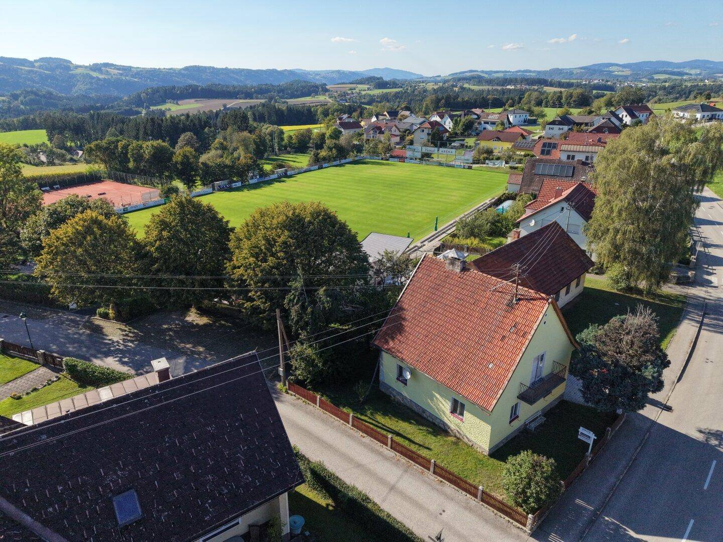 Luftaufnahme des Hauses mit umliegender Landschaft, Sportplatz und Hügeln im Hintergrund.