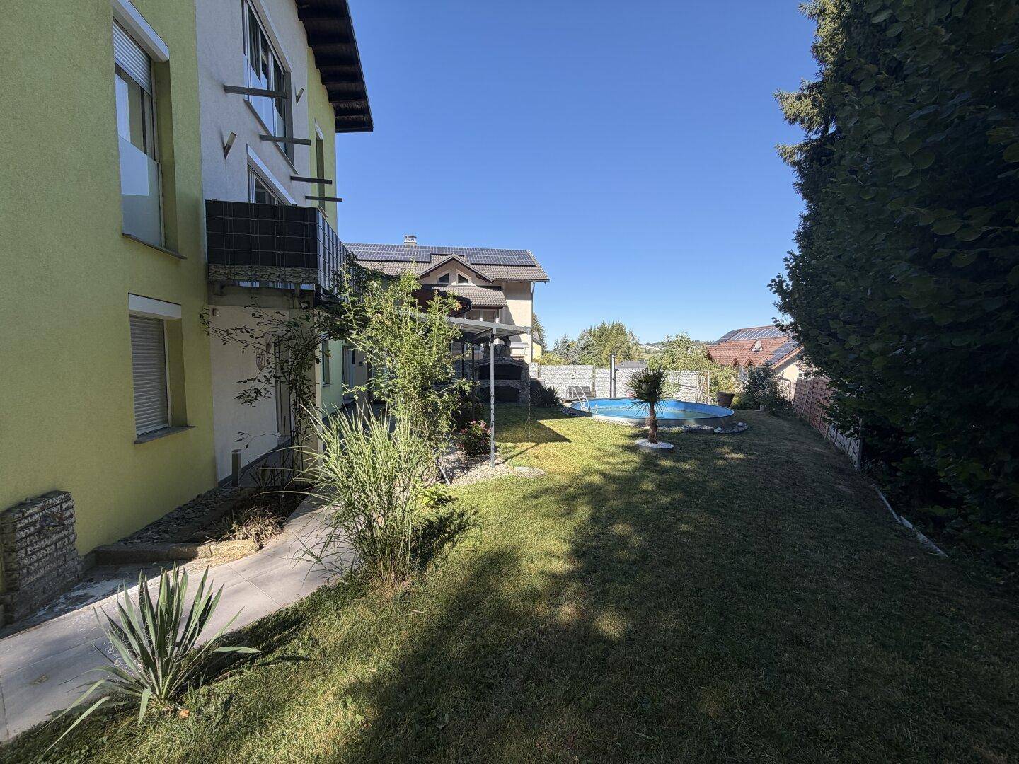 Sonniger Gartenbereich mit Rasenfläche, Pflanzen und Blick auf den Pool im Hintergrund.
