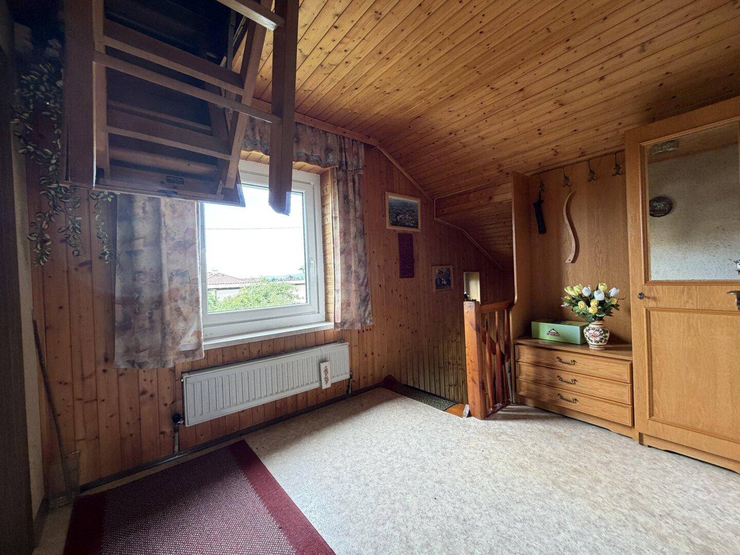 Dachzimmer mit Holzverkleidung, schrägen Wänden und einem Fenster mit Ausblick.