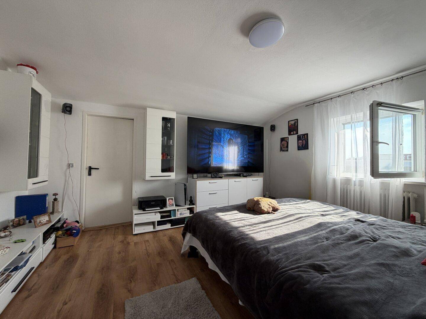 Gemütliches Schlafzimmer mit Holzboden, großem Bett, Wandfernseher und einem Fenster für Tageslicht.