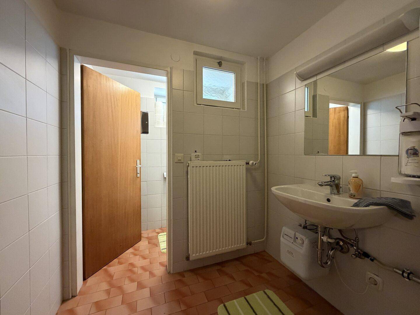 Helles Badezimmer mit weißen Fliesen, Waschbecken, Heizkörper und einem Fenster für natürliche Belüftung.