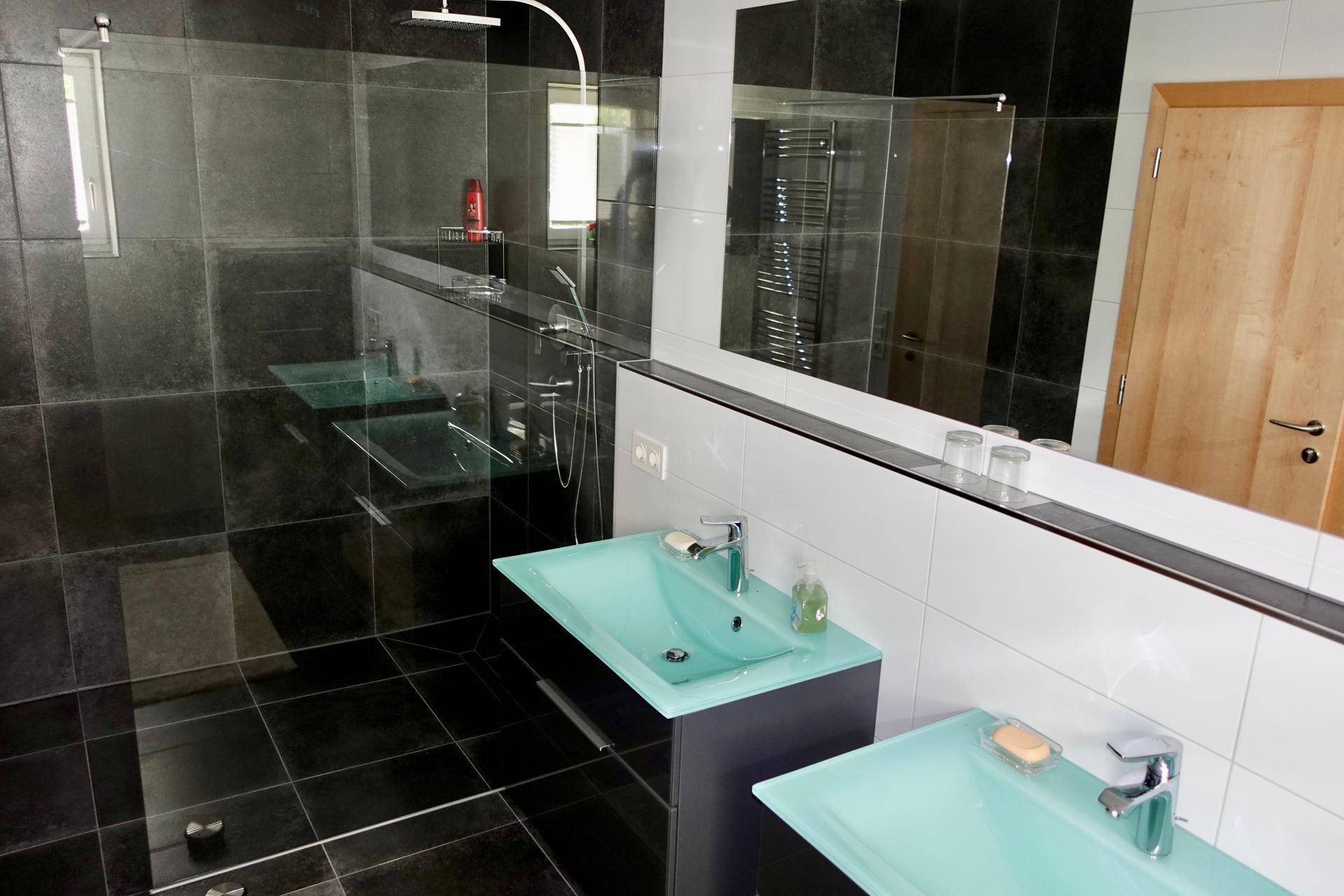 Modernes Badezimmer mit Doppelwaschbecken, großer Dusche und dunklen Fliesen.