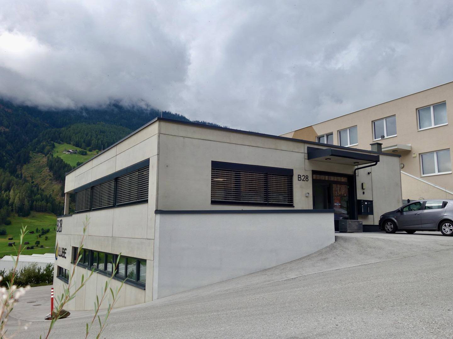 Moderne Architektur eines Geschäftsgebäudes mit klaren Linien und einer Zufahrt, umgeben von grünen Bergen.