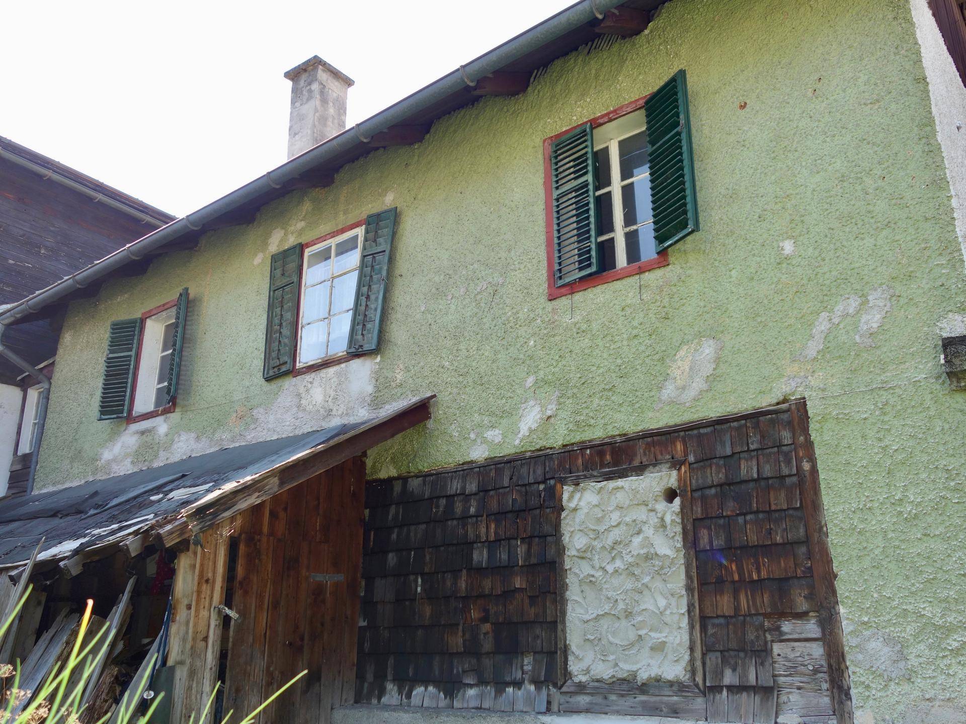 Seitenansicht eines älteren Hauses mit grüner Putzfassade und traditionellen Fensterläden.