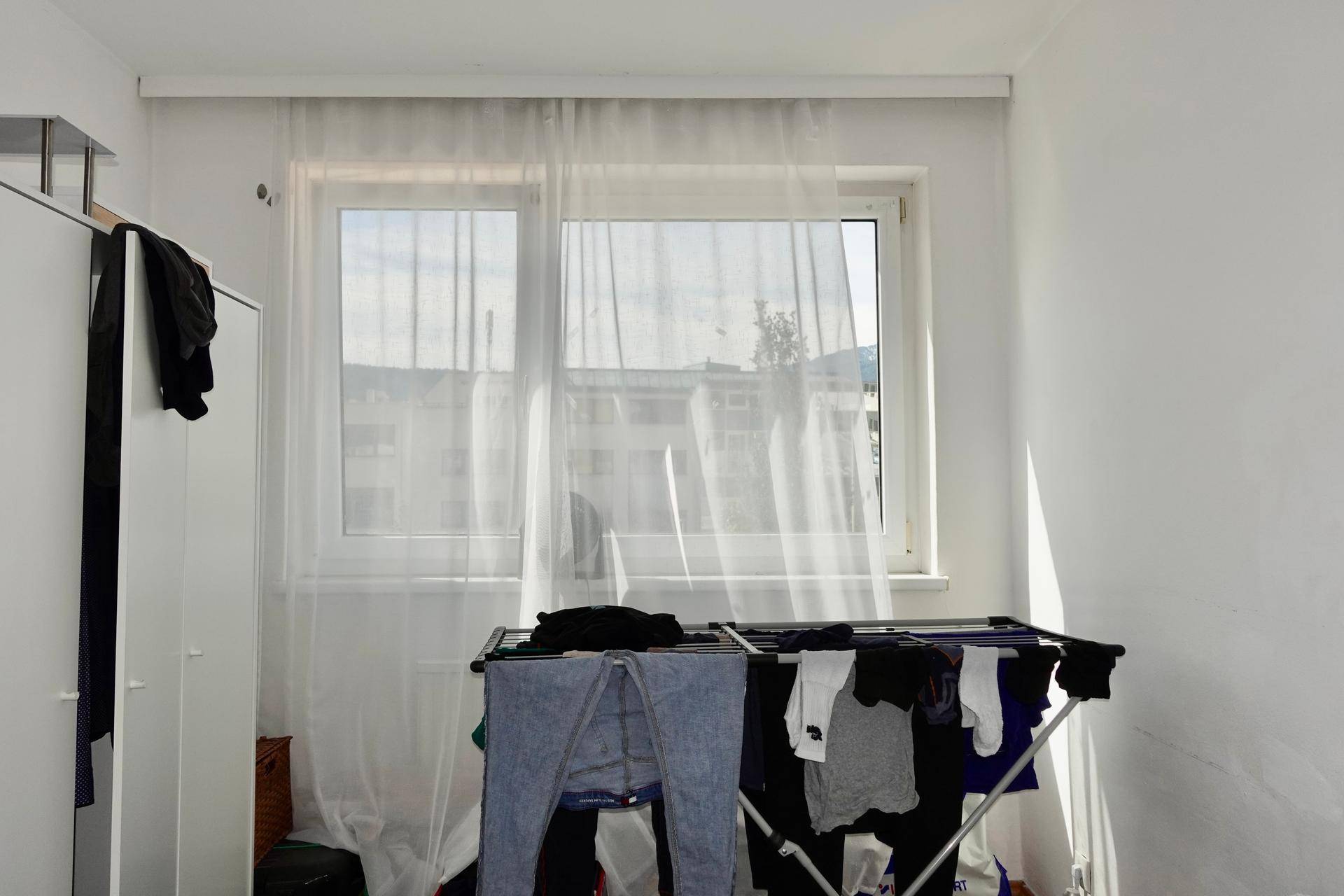 Heller Raum mit Fenster und weißen Vorhängen, Wäscheständer mit Kleidung.