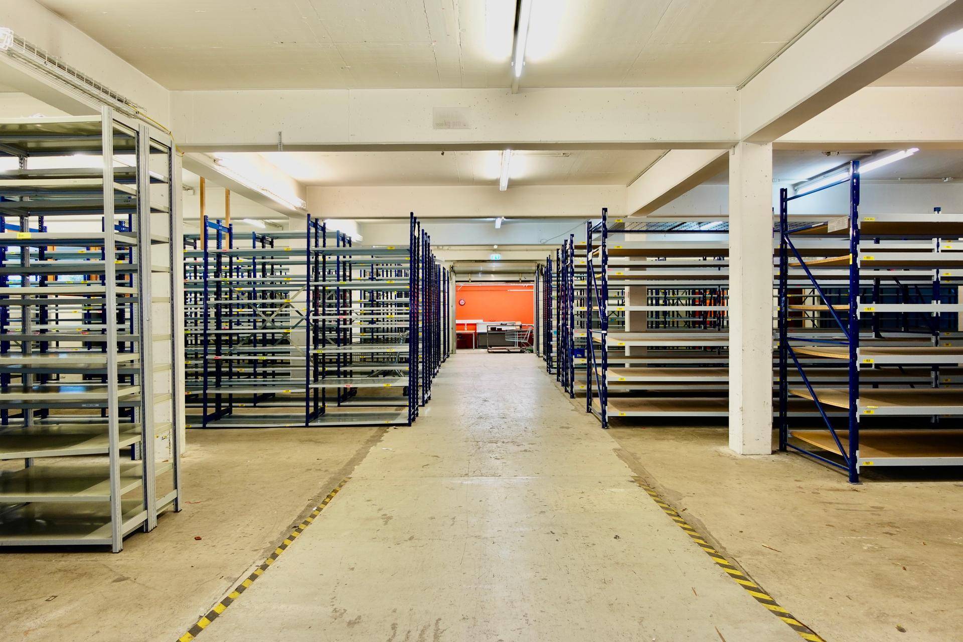 Weitläufige Lagerhalle mit leeren Regalsystemen und gelb-schwarzen Bodenmarkierungen.