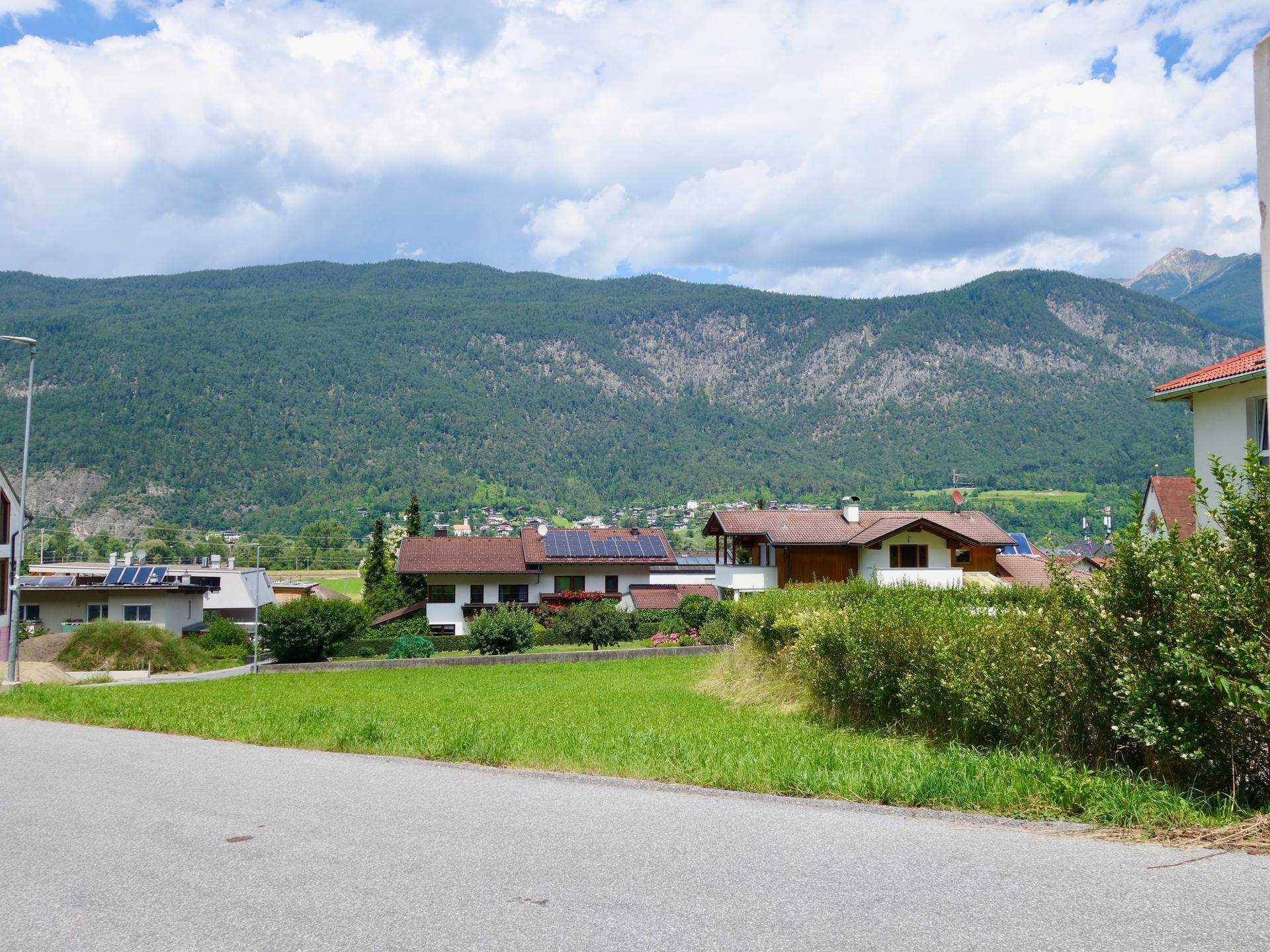 Wohngebiet mit Häusern und Solaranlagen auf den Dächern, umgeben von grünen Hügeln und Bergen unter blauem Himmel.
