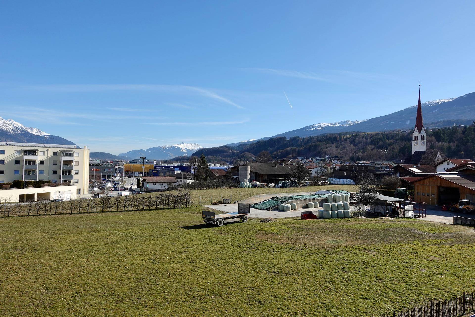 Weitläufiger Blick über grüne Felder auf eine Ortschaft mit Kirche und schneebedeckten Bergen im Hintergrund.