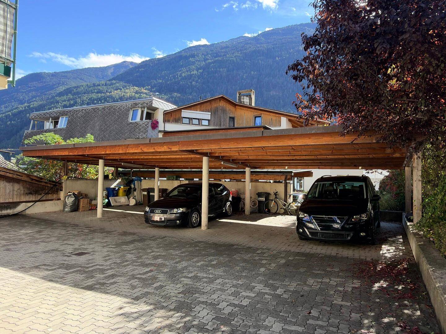 Überdachter Parkplatz mit mehreren Stellplätzen und Autos, umgeben von Bäumen und Bergen.