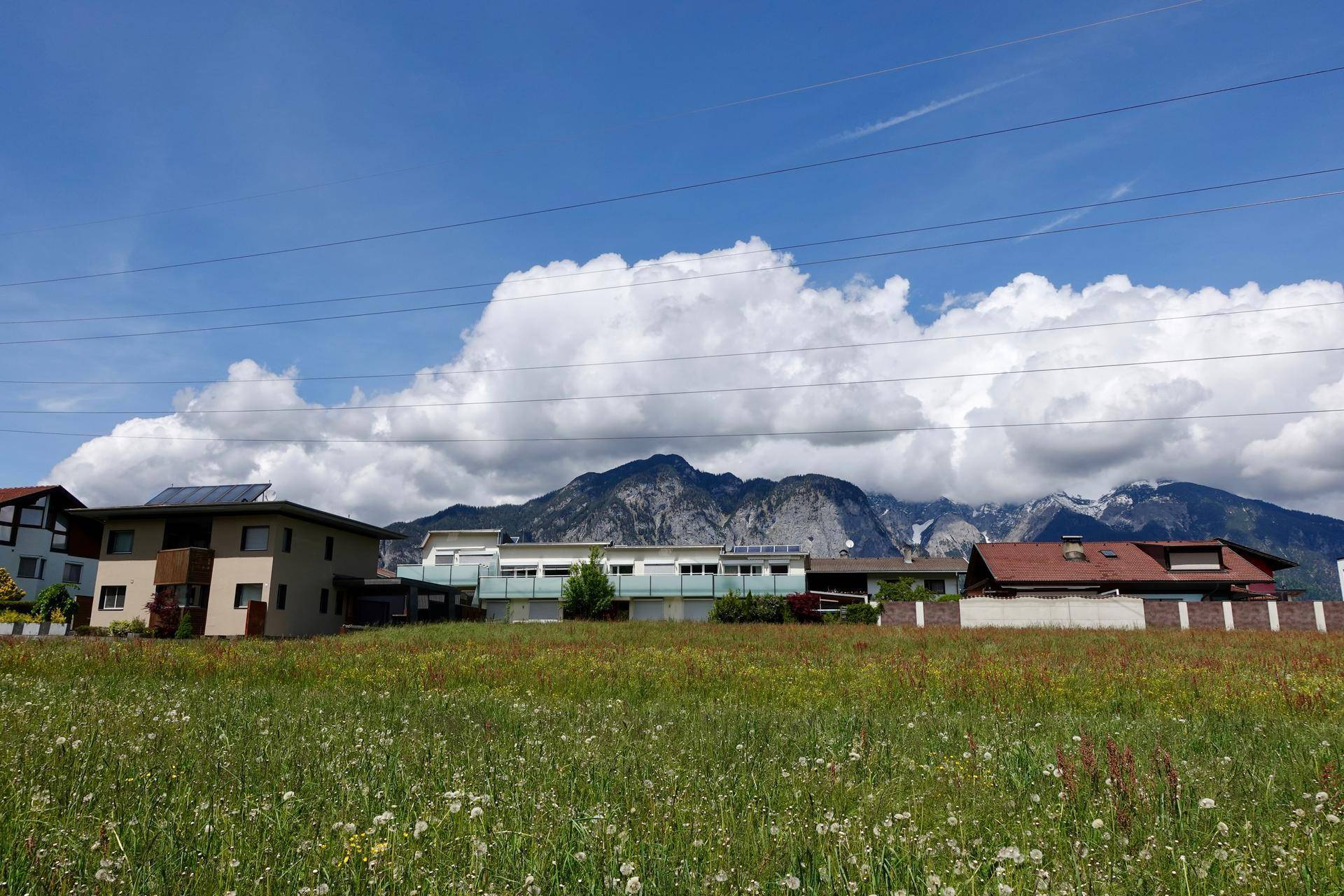 Weitläufige Landschaft mit Häusern, grünen Wiesen und majestätischen Bergen unter bewölktem Himmel.
