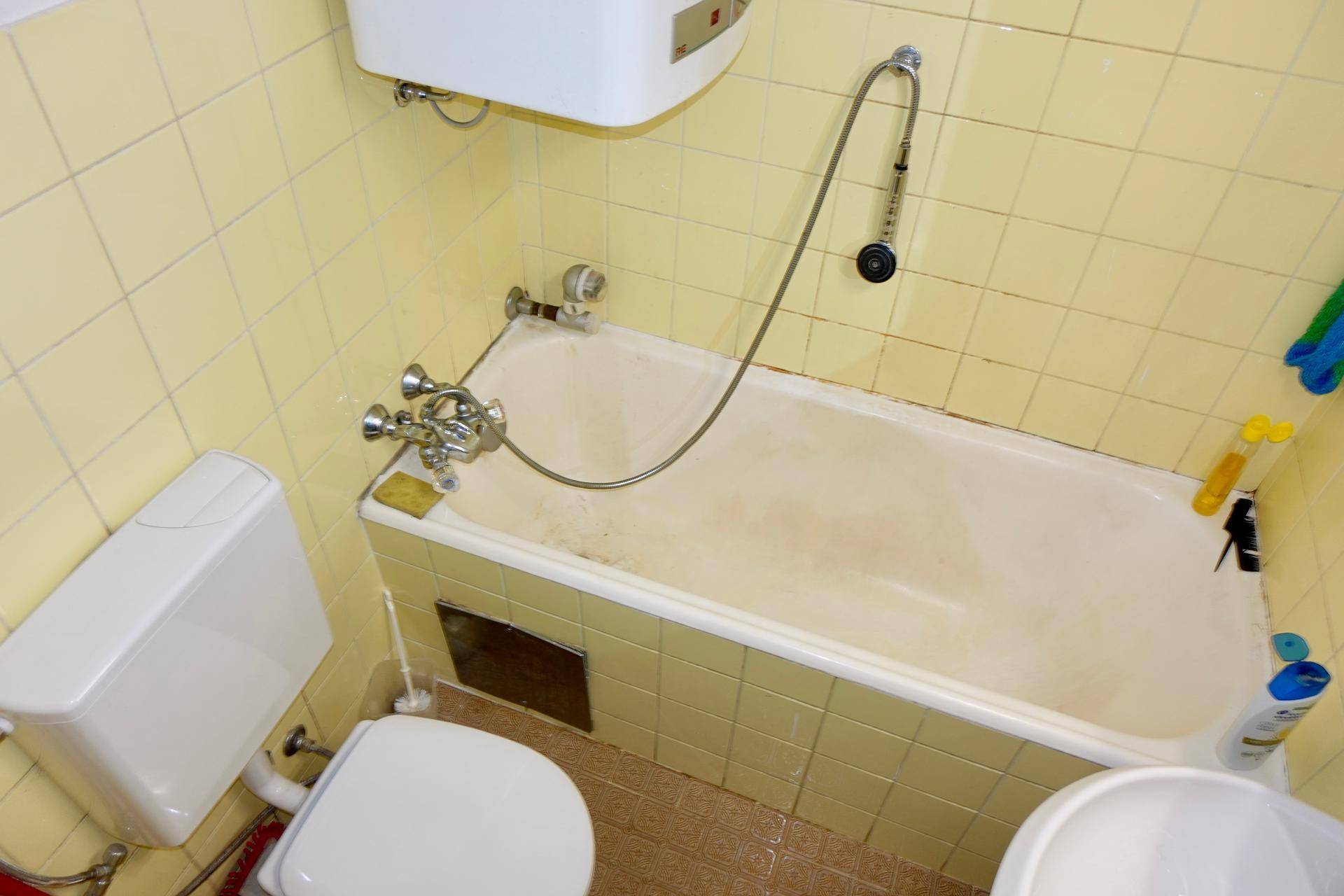 Funktionales Badezimmer mit gelben Fliesen, Badewanne mit Duschfunktion und Toilette im Originalzustand.
