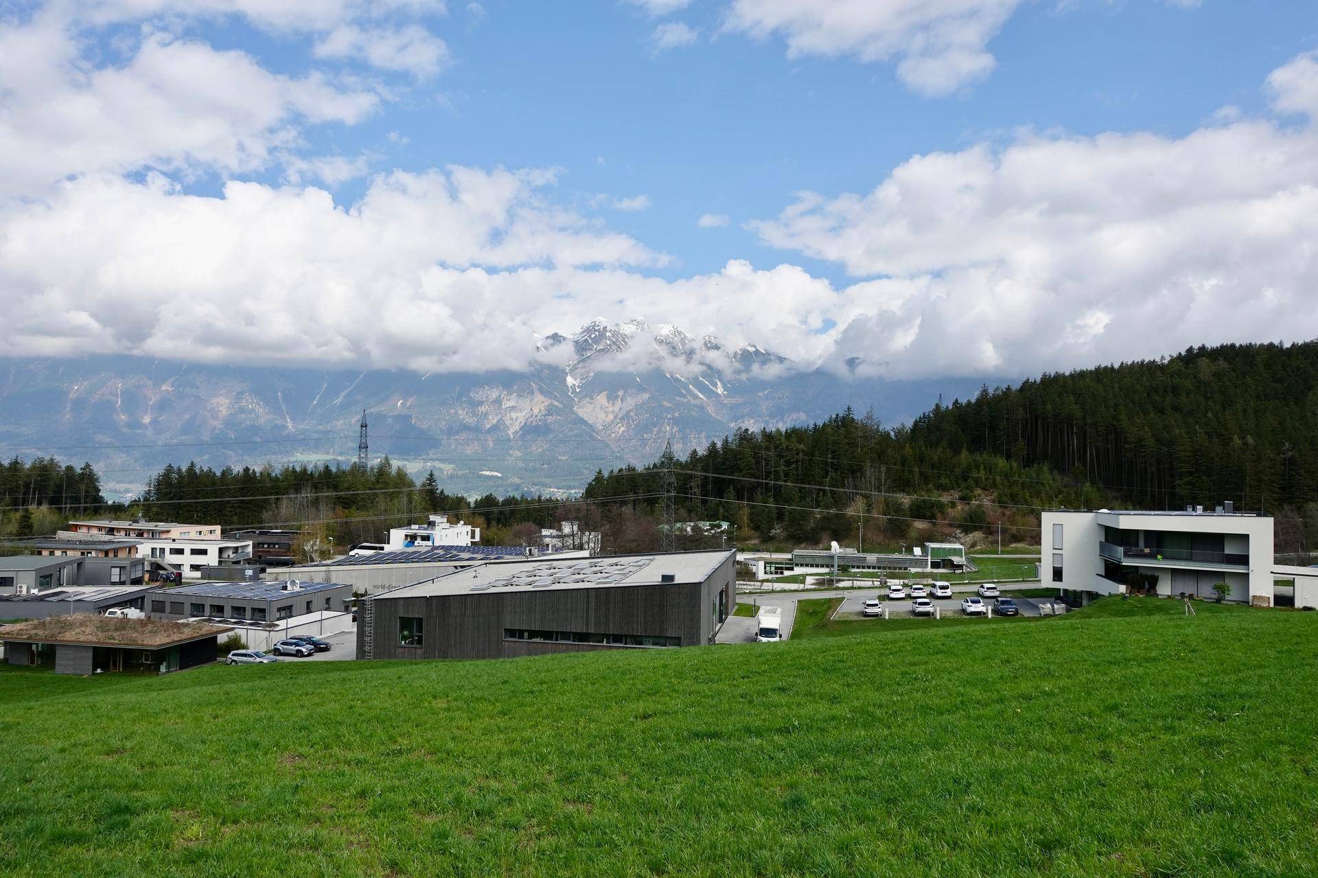 Panoramablick über eine grüne Landschaft mit modernen Gebäuden, Parkplätzen und majestätischen Bergen im Hintergrund.