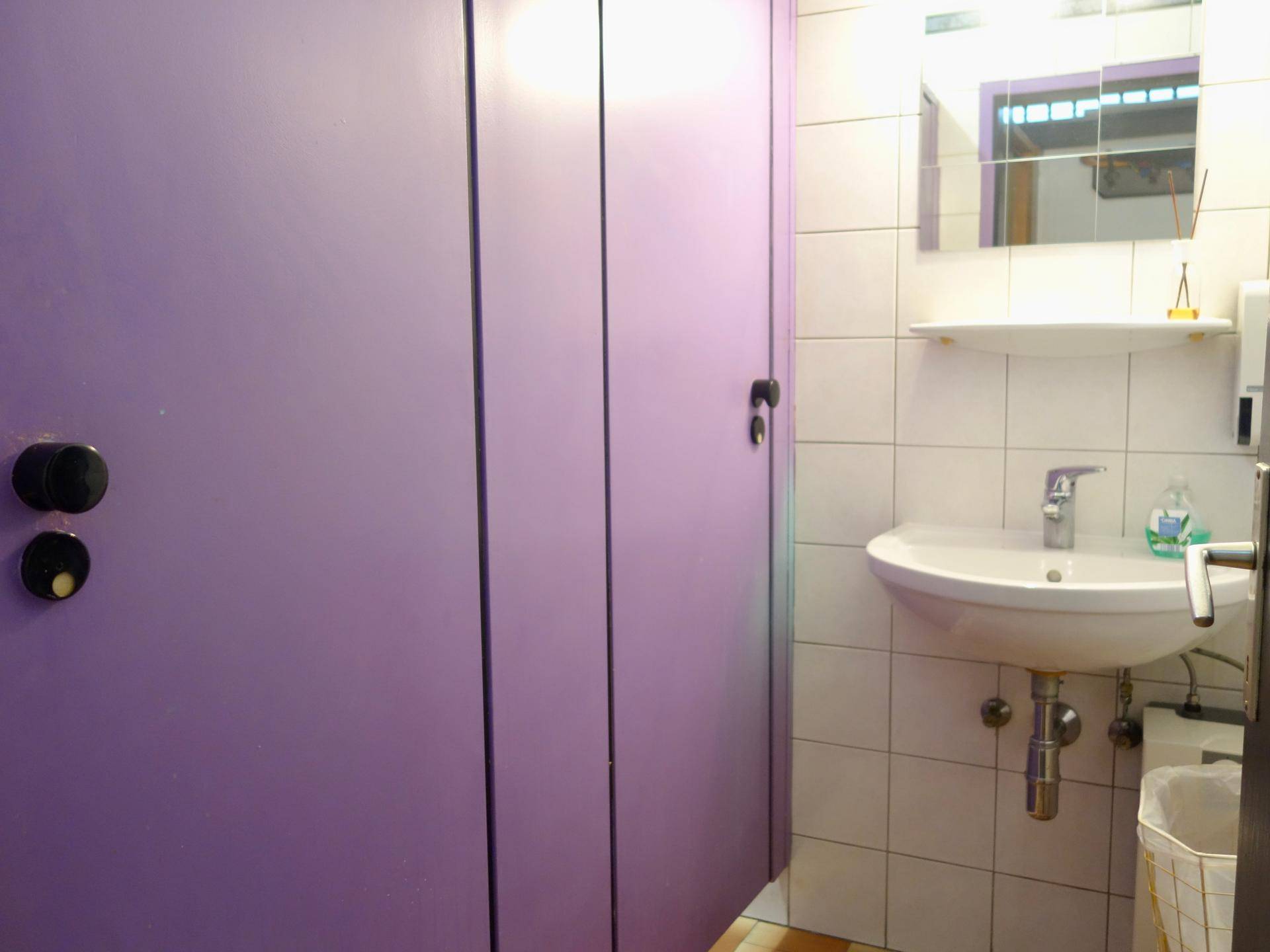 Toilettenbereich mit lila Kabinentüren, einem Waschbecken und weißen Wandfliesen.