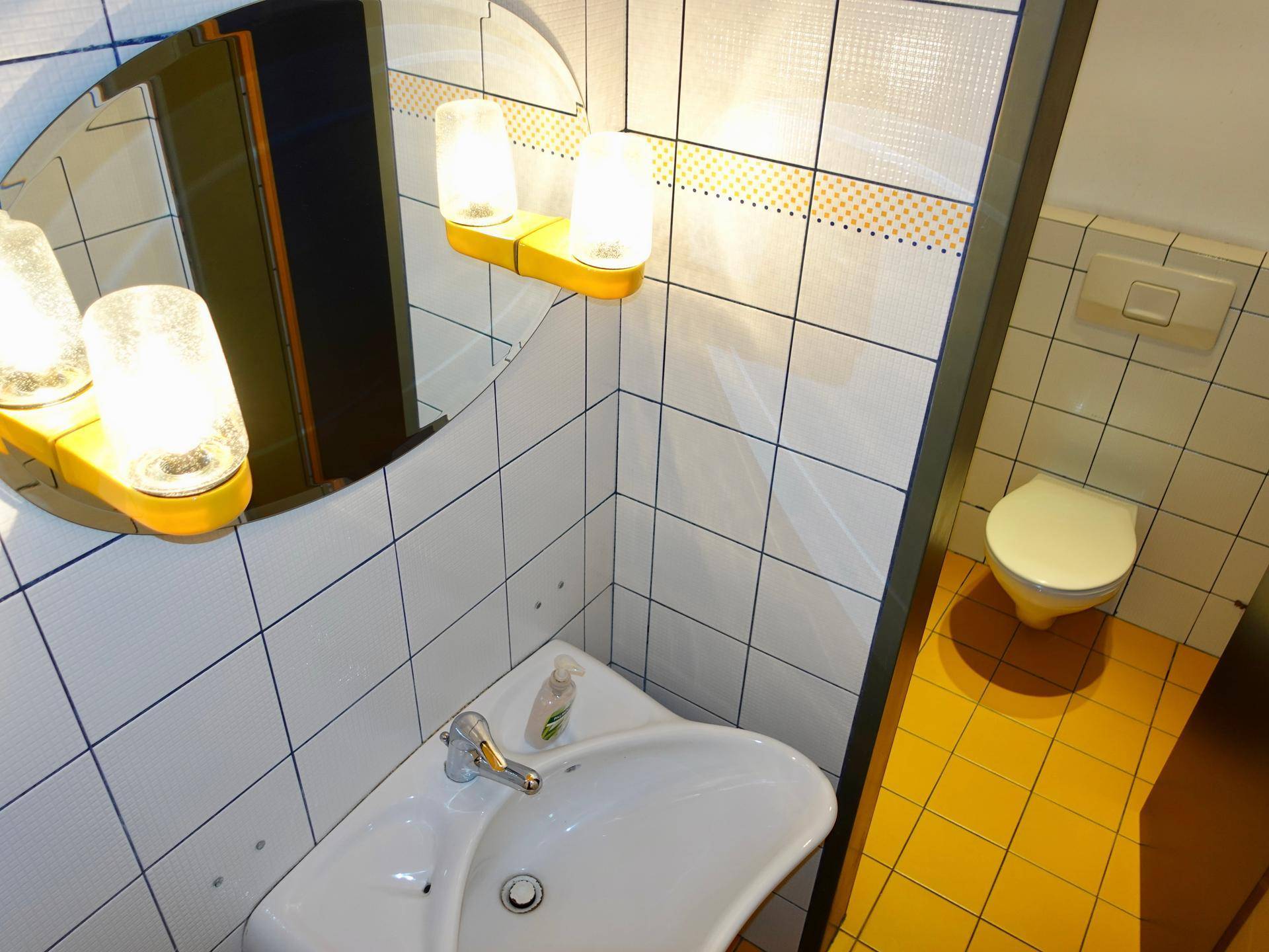 Kleines Badezimmer mit gelben Fliesen, Waschbecken, Toilette und Spiegel mit Beleuchtung.