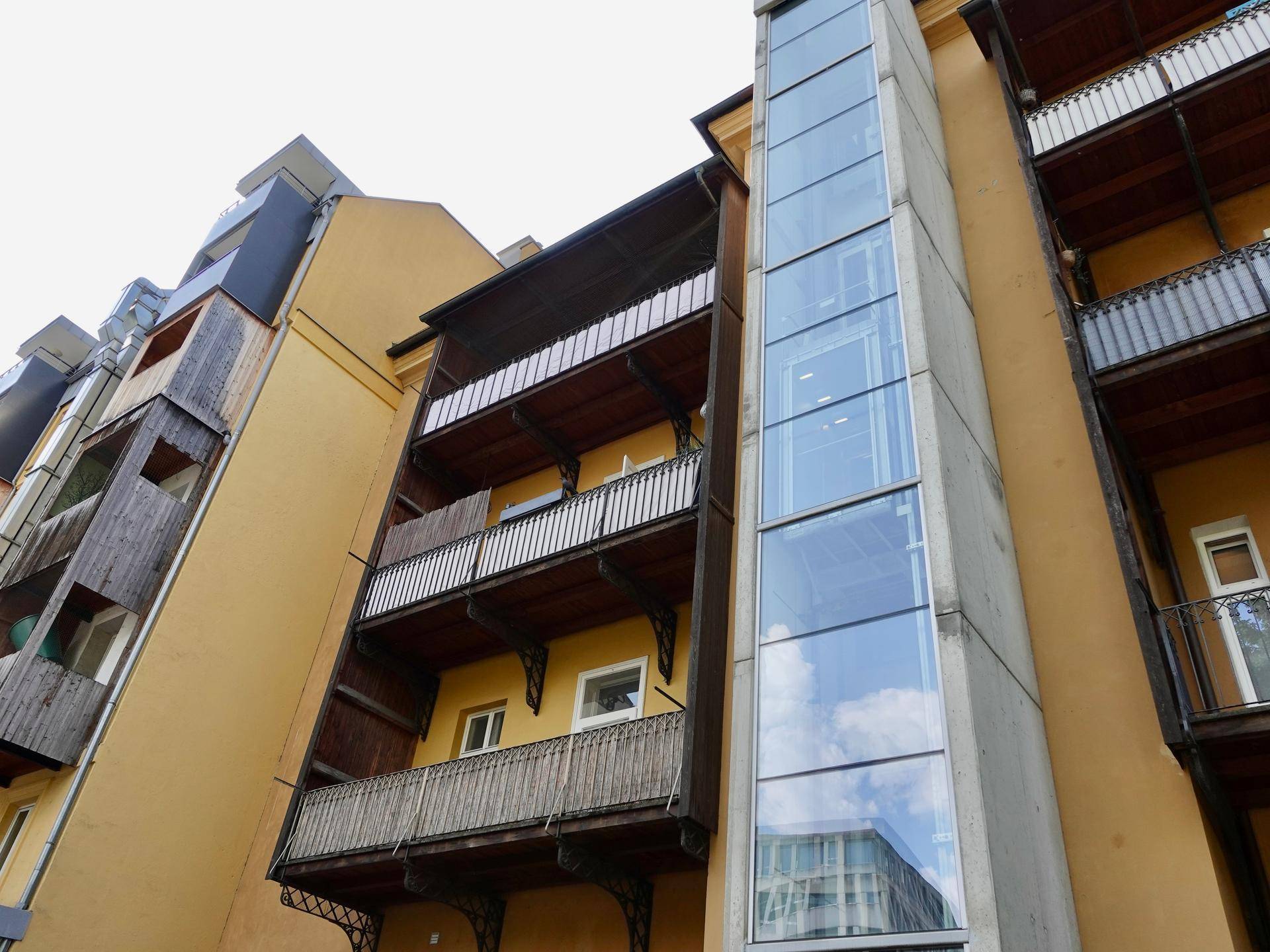 Außenansicht eines Gebäudes mit mehreren Balkonen und einem modernen Glasaufzug an der Fassade.