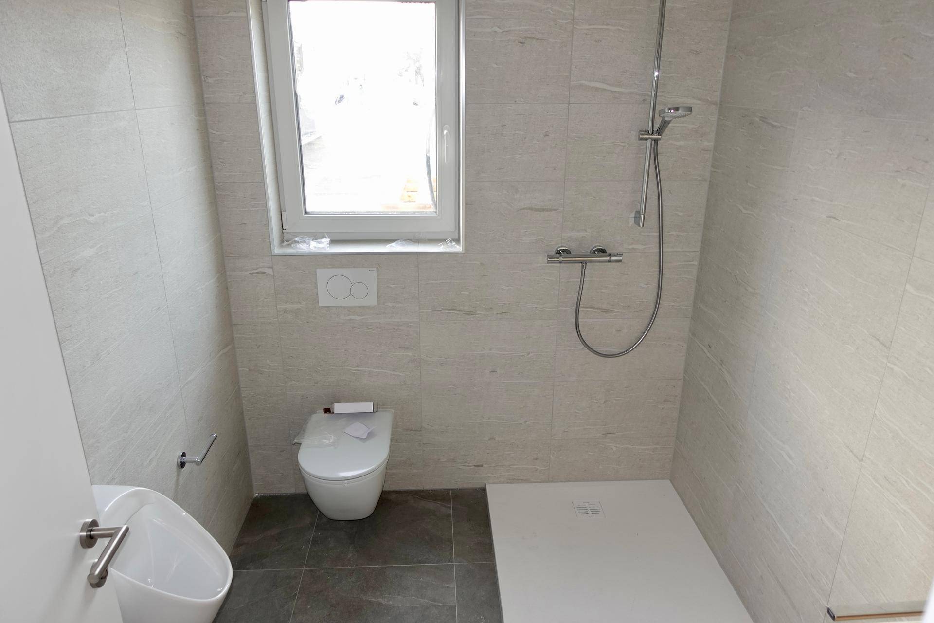 Helles Badezimmer mit Dusche, Toilette, Urinal und einem Fenster für natürliche Belüftung.