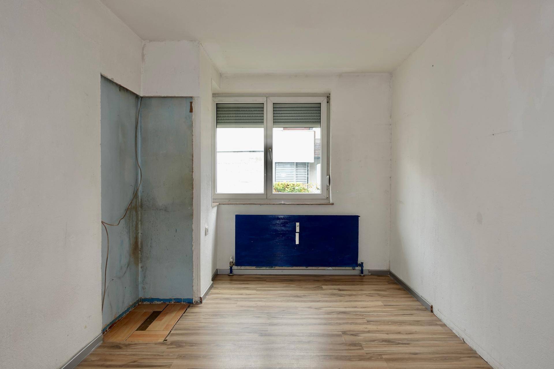 Leerer Raum mit Laminatboden, Fenster und einem blauen Heizkörper, bereit zur Renovierung.