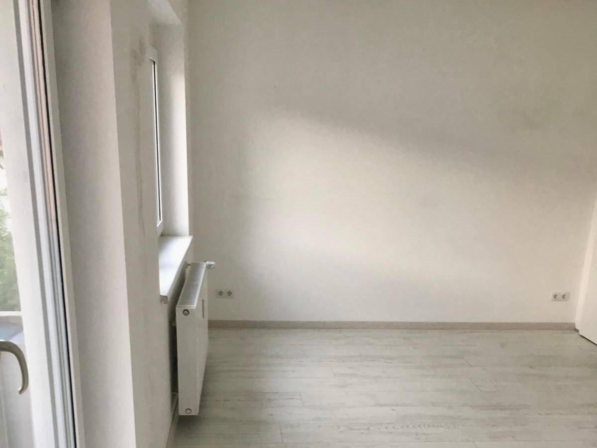 Leerer Raum mit hellen Wänden, Laminatboden und einem Fenster mit Heizkörper darunter.