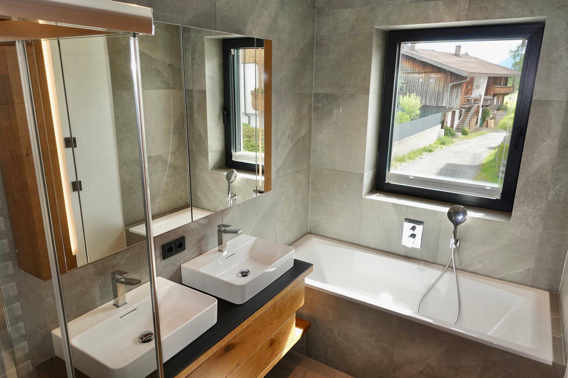 Badezimmer mit Badewanne, Doppelwaschbecken, Spiegelschrank und Fenster für Tageslicht.