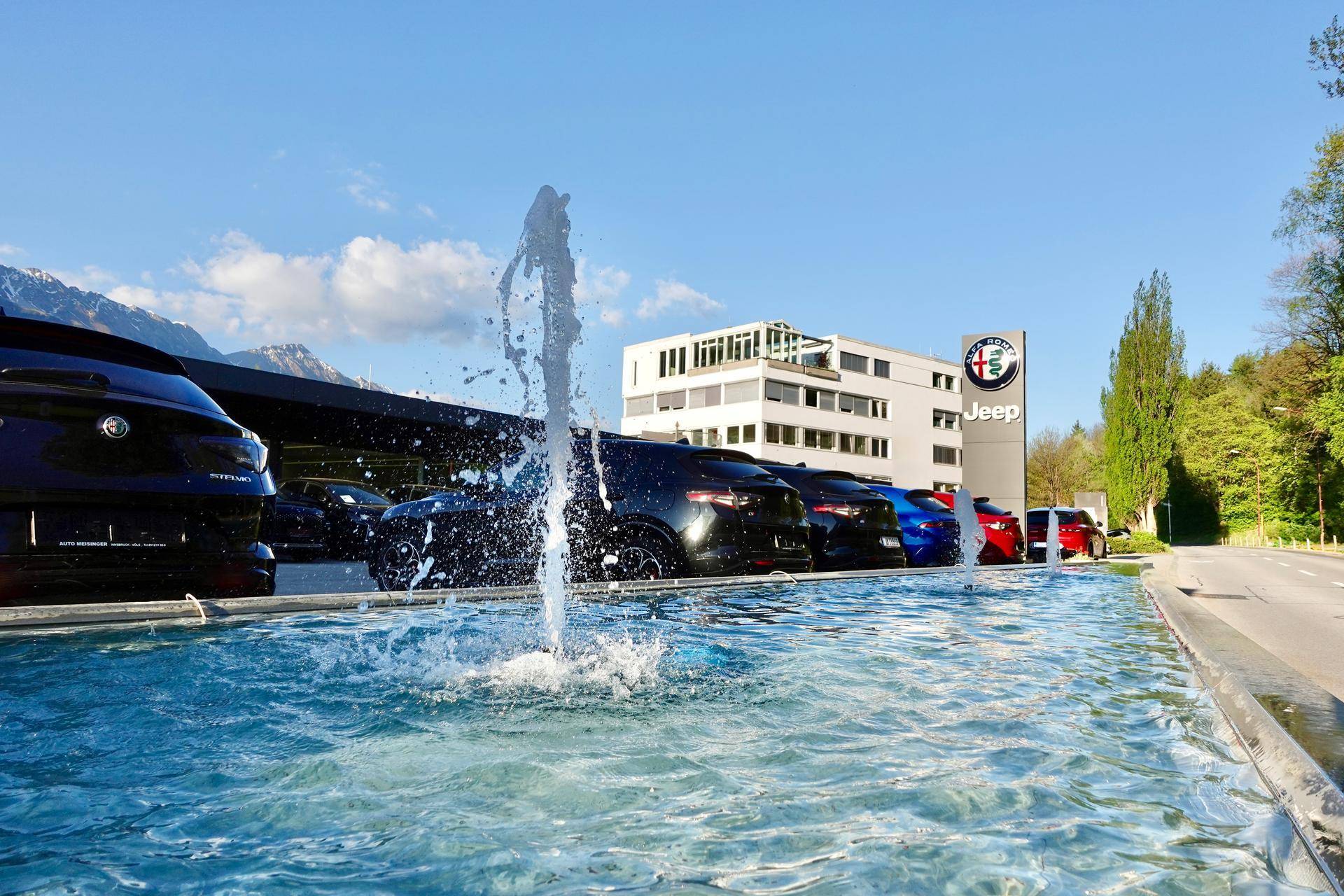 Außenansicht eines Geschäftsgebäudes mit Springbrunnen und geparkten Autos unter blauem Himmel.