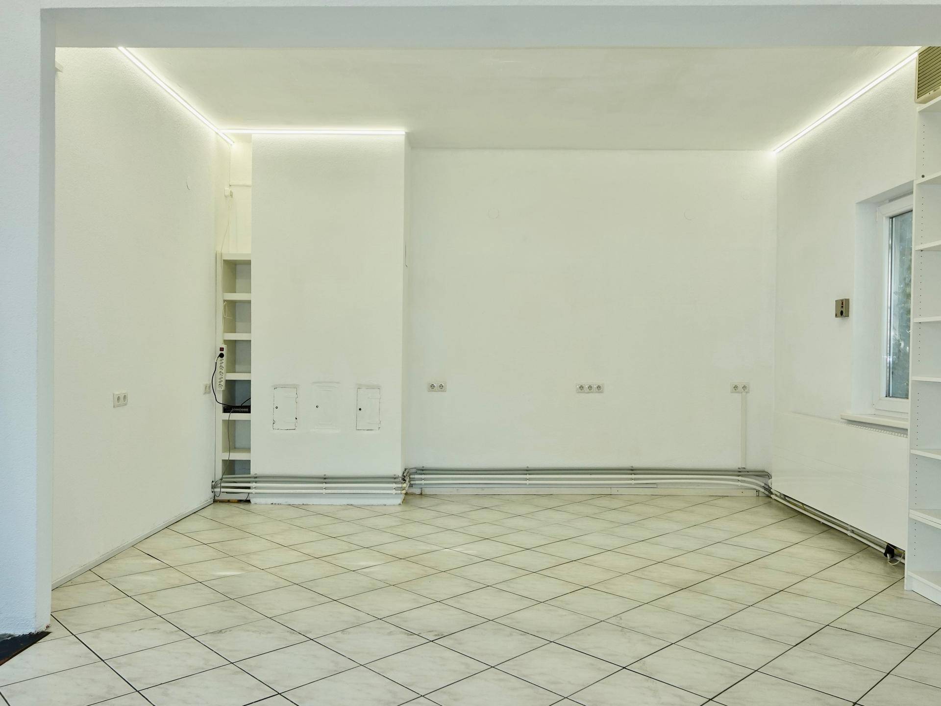 Teilansicht eines leeren Gewerberaums mit weißen Wänden, sichtbaren Heizungsrohren und zahlreichen Steckdosen.
