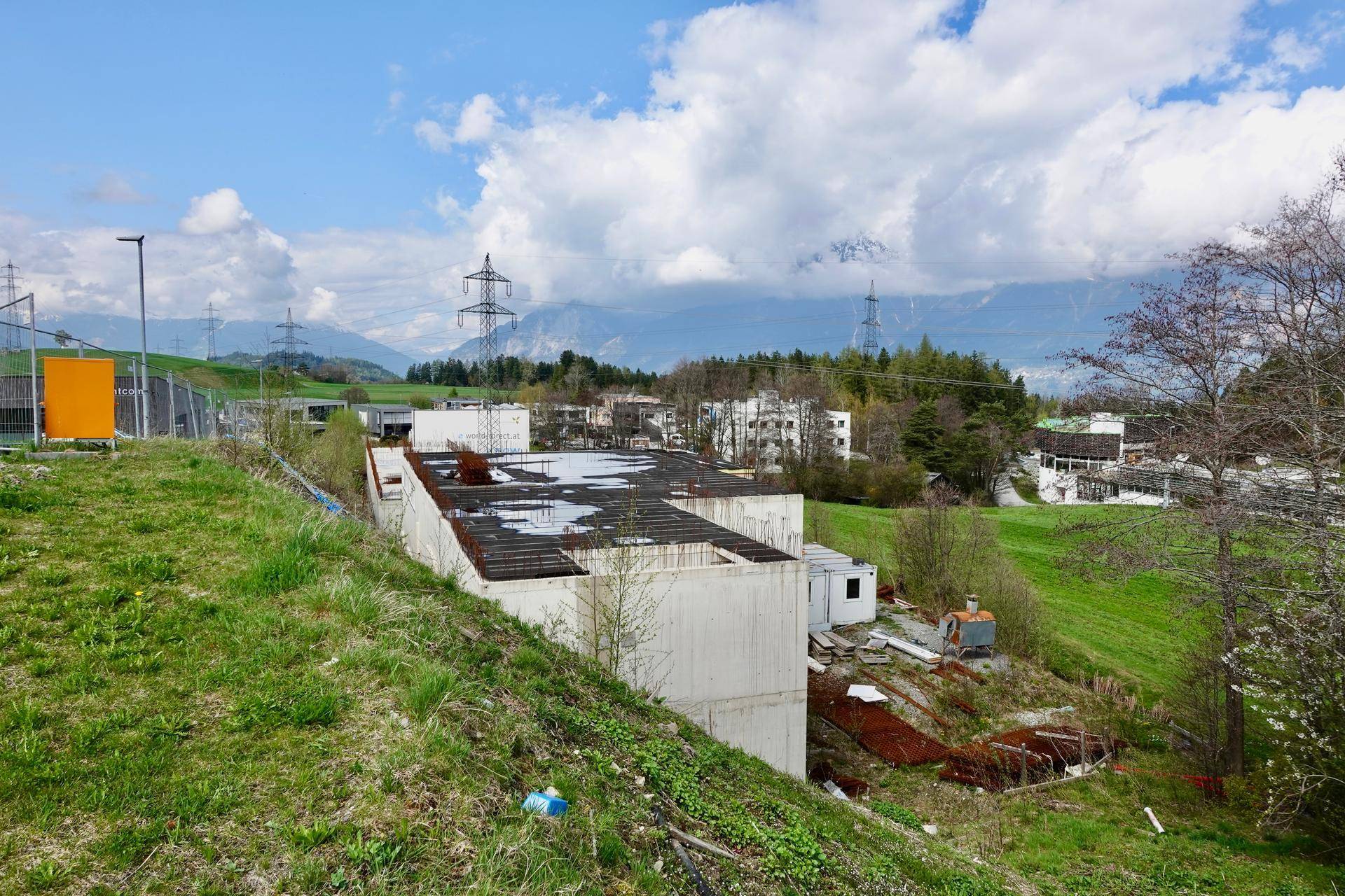 Blick von einem Grashang auf eine Baustelle mit Betonstrukturen und Bewehrungsstahl, umgeben von grüner Landschaft und Bergen.