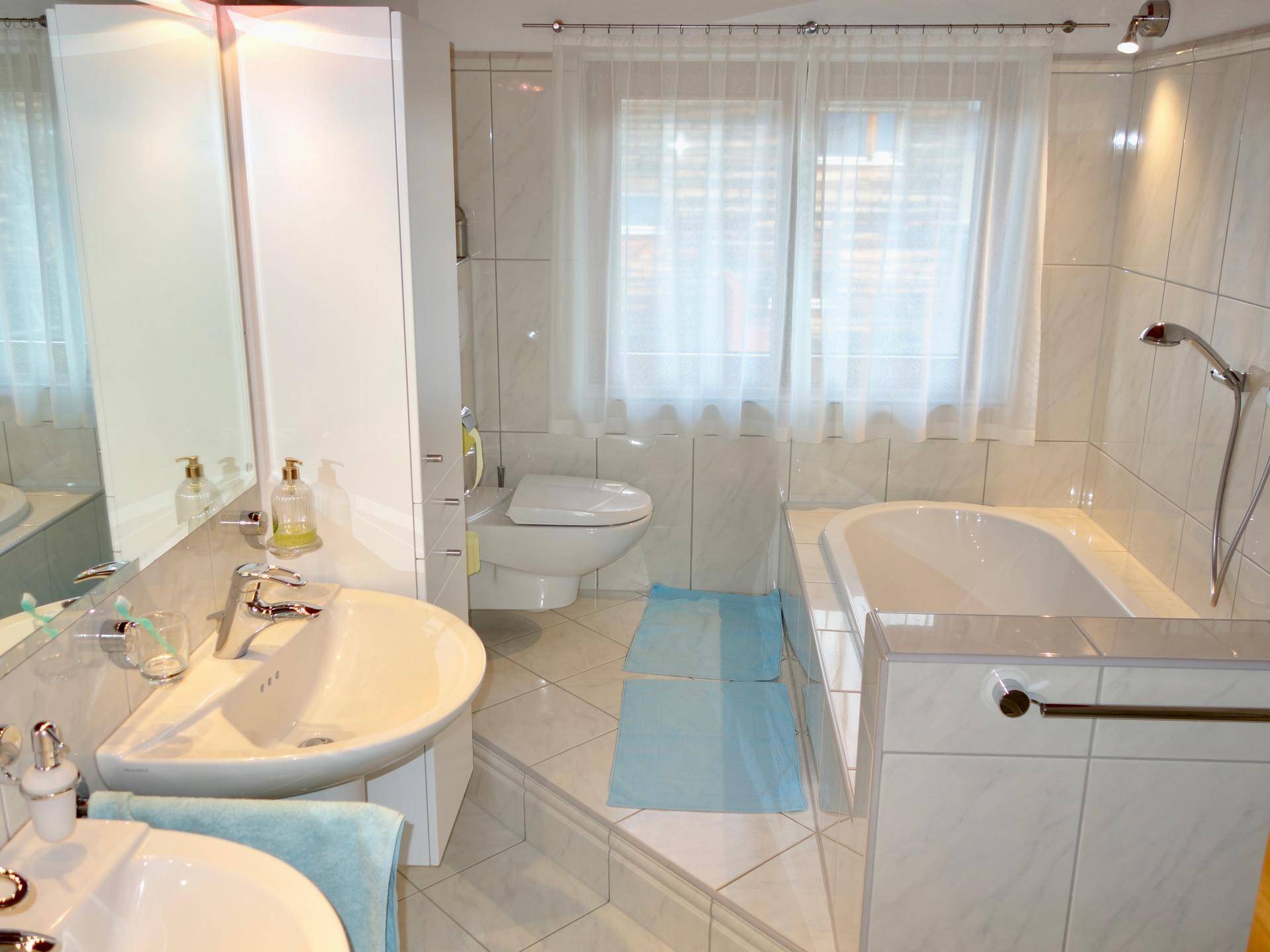 Helles Badezimmer mit Badewanne, Doppelwaschbecken, WC und Fenster.