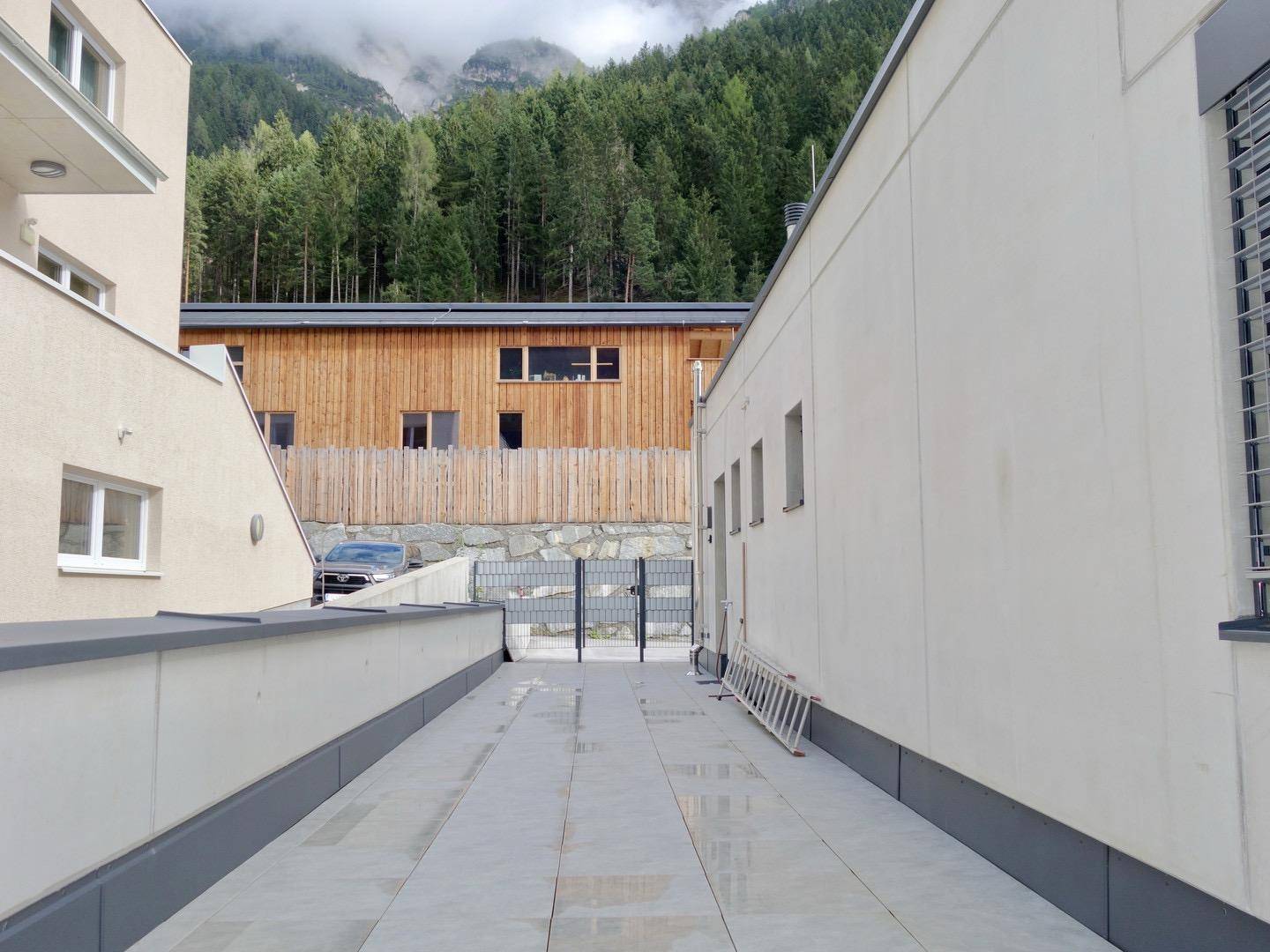 Gepflasterter Außenbereich zwischen Gebäuden mit Holzzaun und Blick auf bewaldete Berghänge.