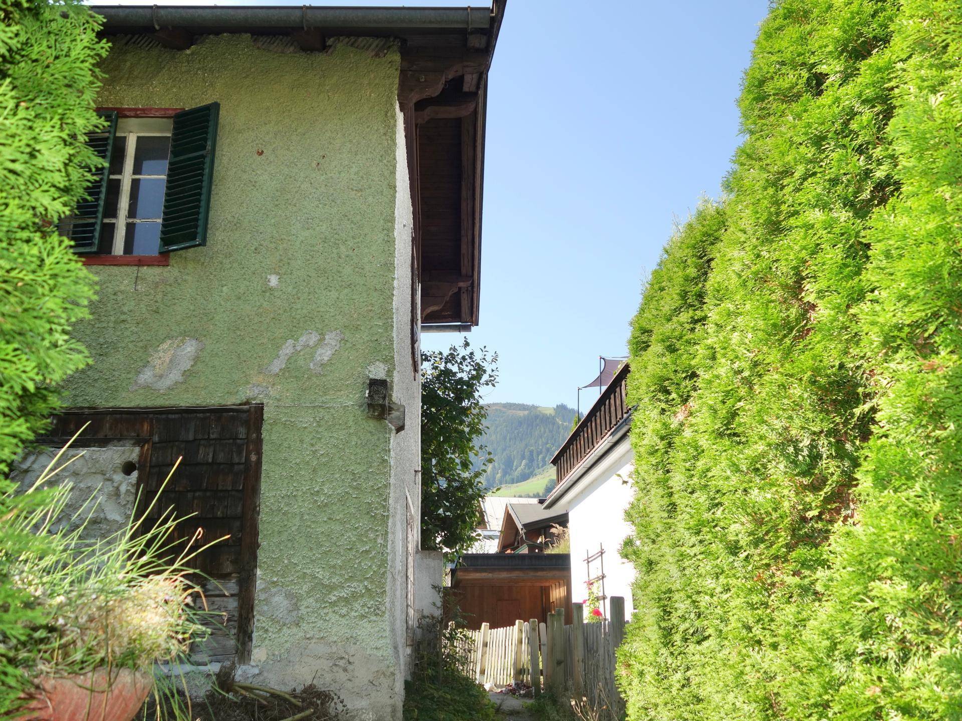 Schmaler Durchgang zwischen Gebäuden mit hoher Hecke und Blick auf die umliegende Berglandschaft.