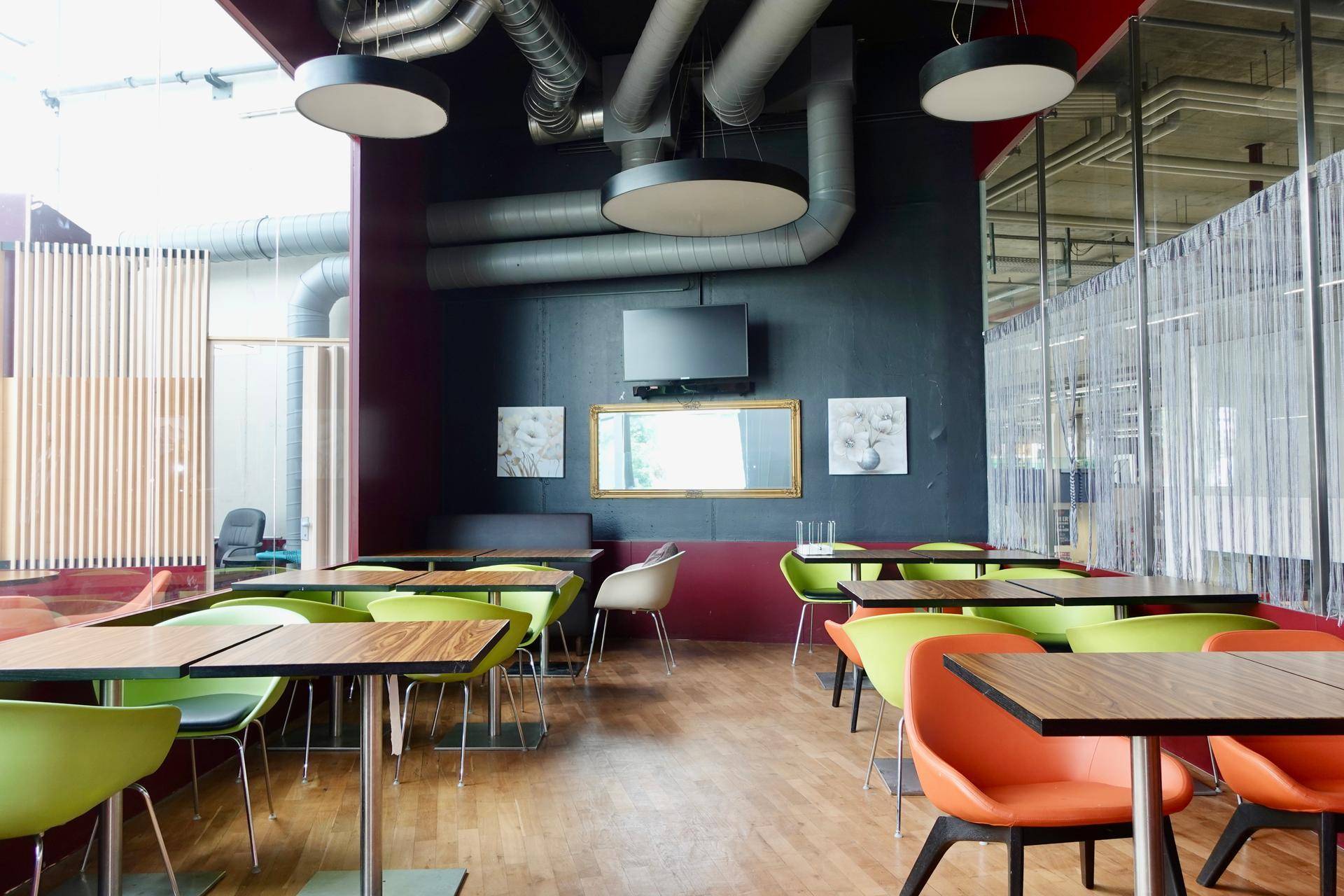 Gemütlicher Restaurantbereich mit farbigen Stühlen, Holzboden und sichtbaren Lüftungsrohren an der Decke.