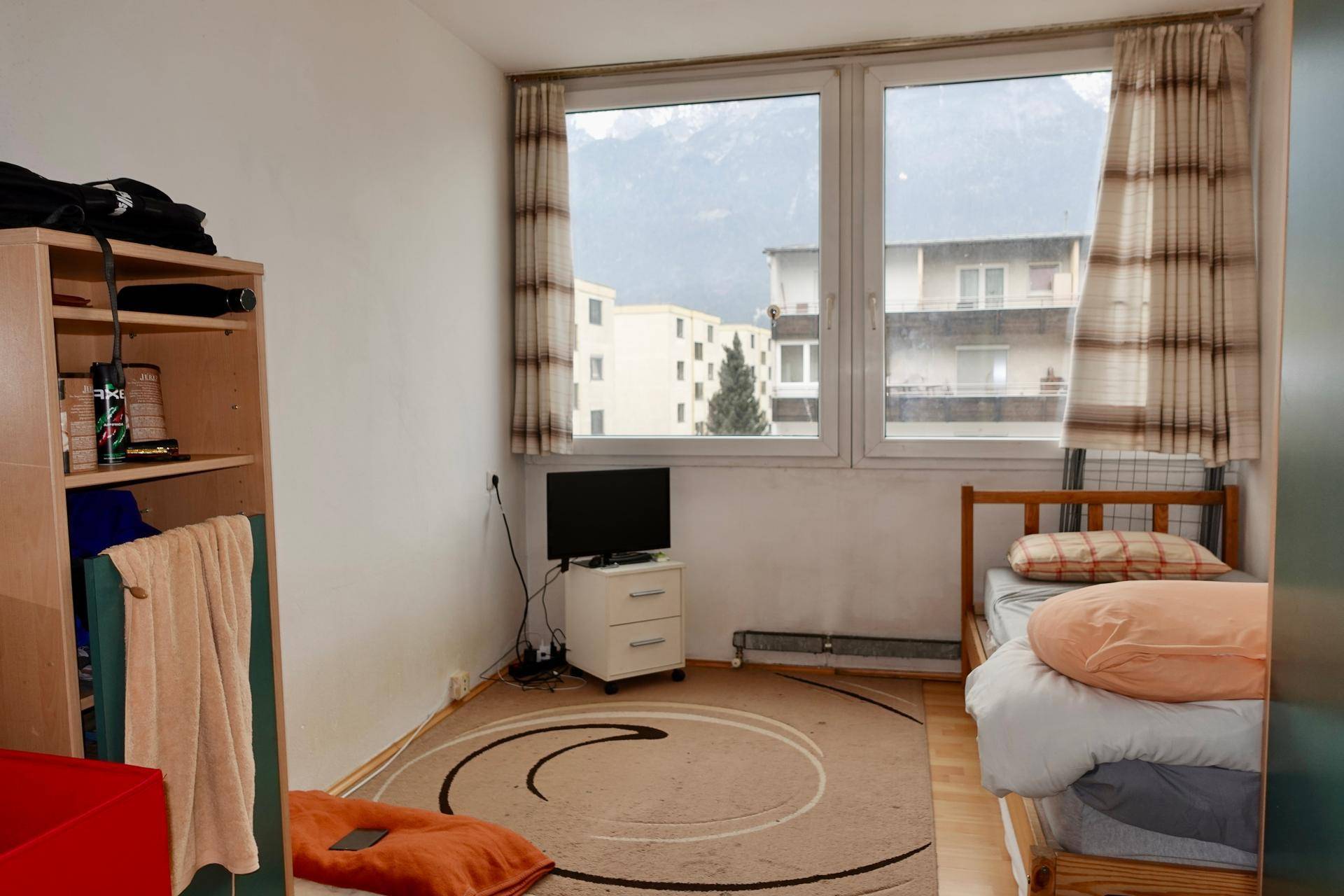 Einfaches Schlafzimmer mit Einzelbett, Teppichboden und Blick auf die umliegenden Gebäude und Berge.