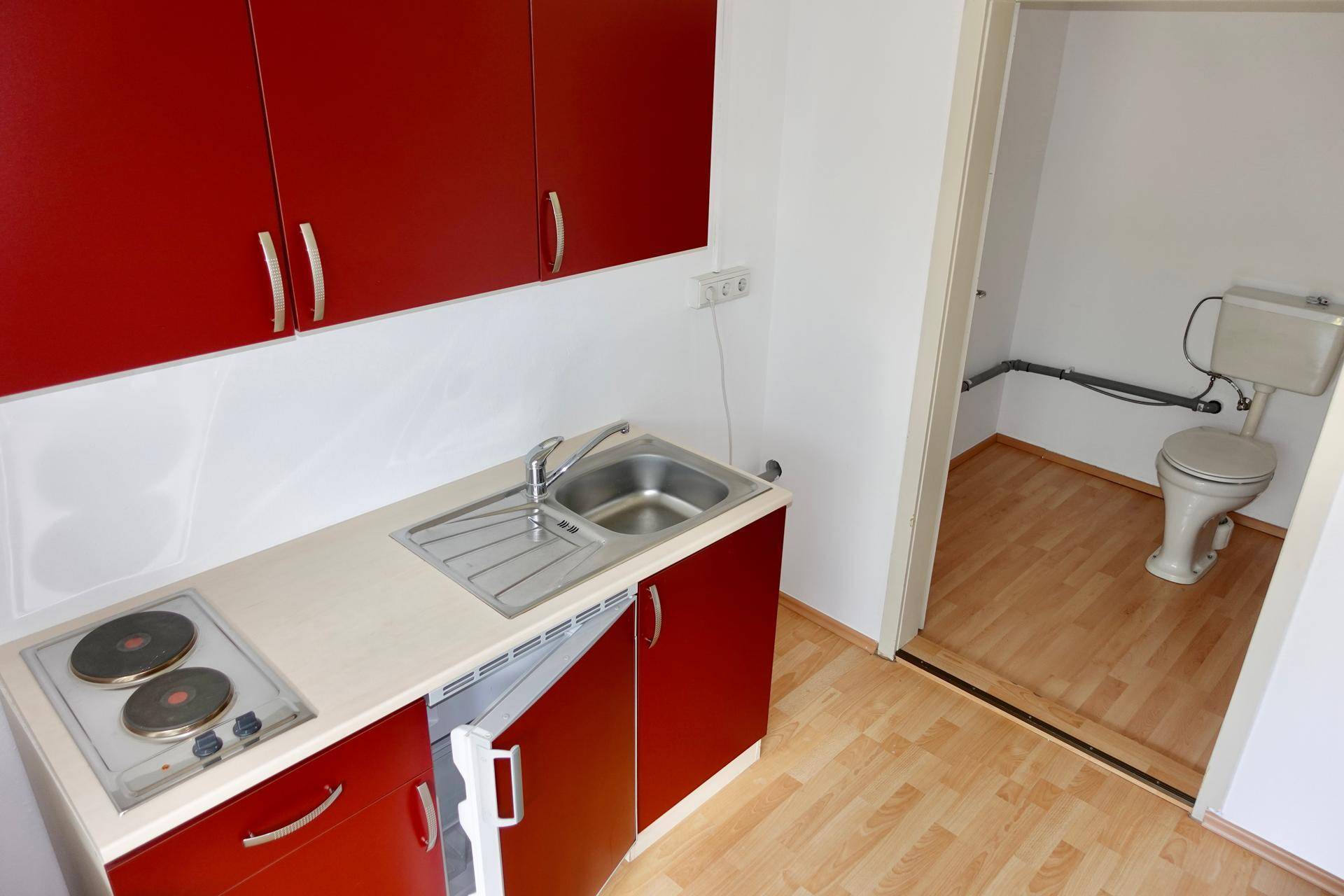 Kompakte Küchenzeile mit roten Schränken, Spüle und Kochplatten, angrenzend an eine kleine Toilette.