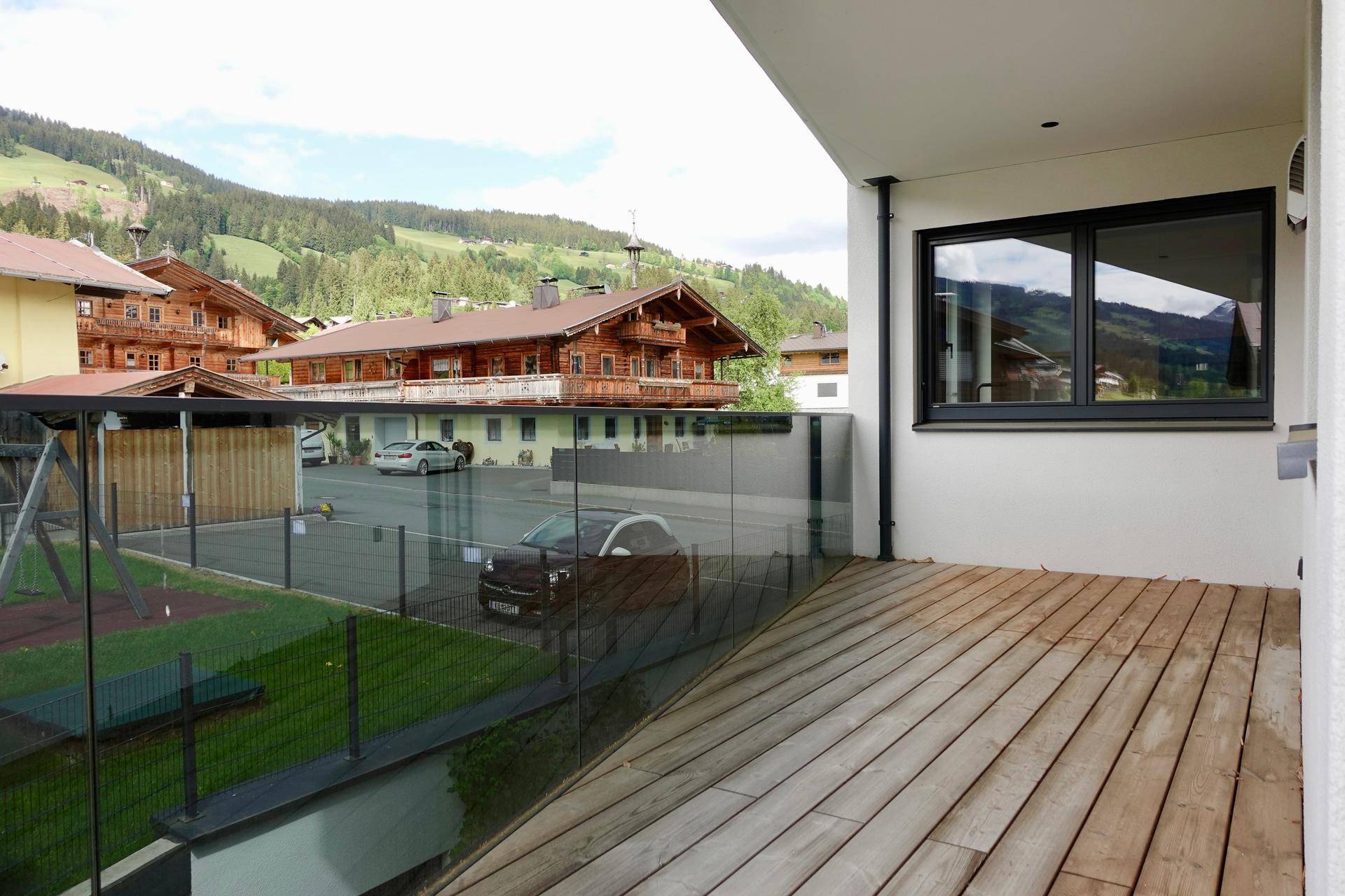 Balkon mit Holzboden, Glasbrüstung und Blick auf die umliegenden Häuser und Berge.