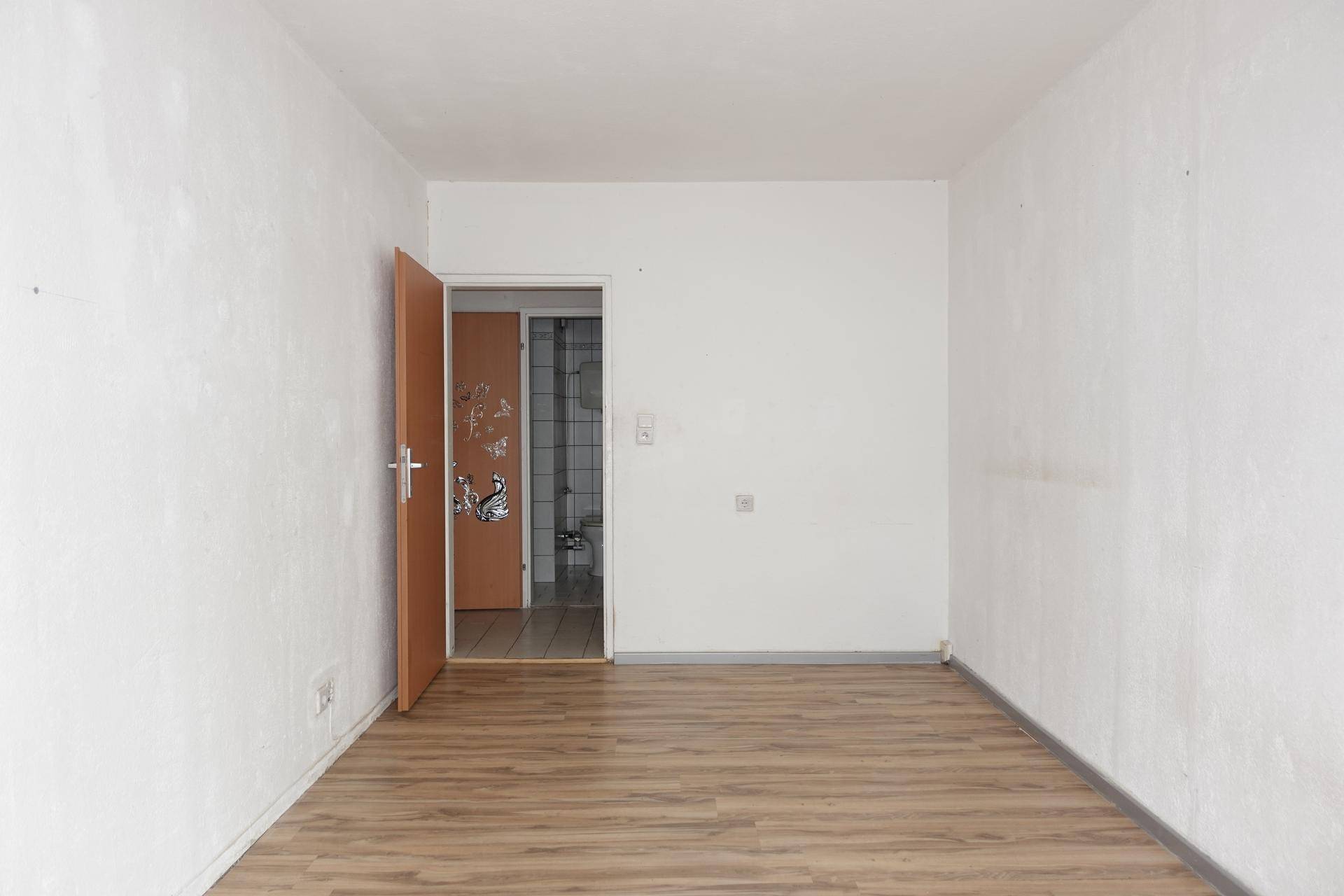 Leerer Raum mit Laminatboden und weiß gestrichenen Wänden, eine Tür führt ins Badezimmer.
