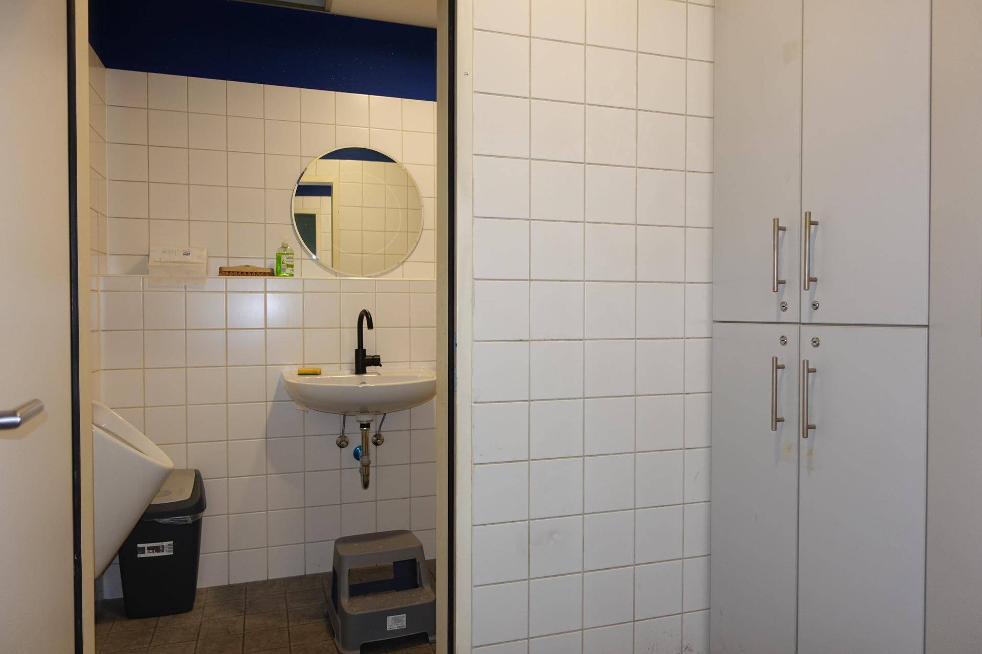 Öffentliche Toilette mit Waschbecken, rundem Spiegel, Urinal und weißen Fliesen an den Wänden.