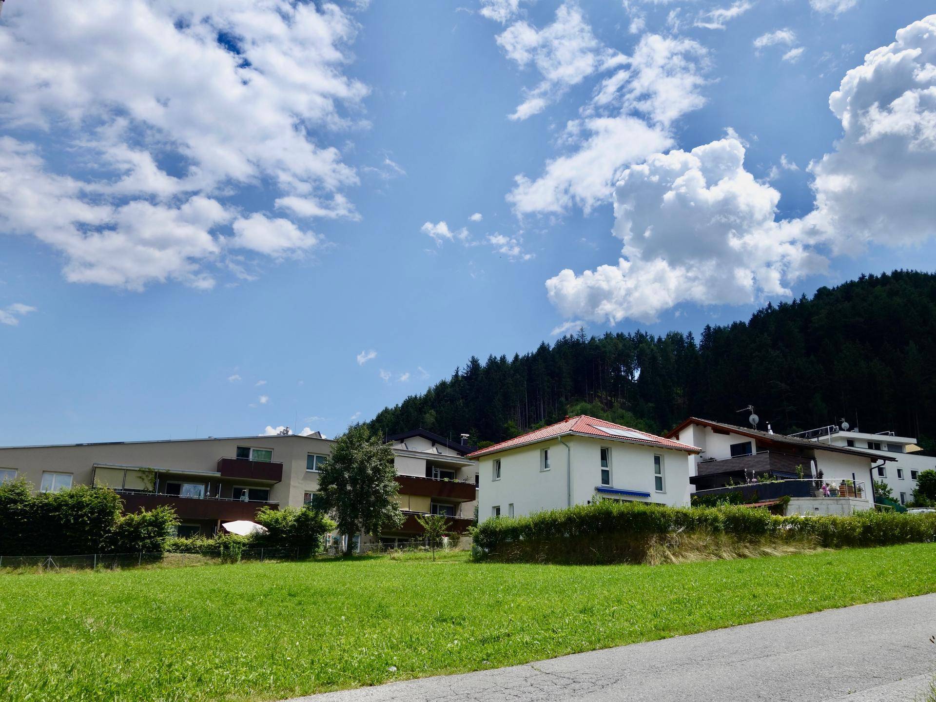 Moderne und traditionelle Häuser am Fuße eines bewaldeten Hügels unter einem strahlend blauen Himmel mit Wolken.