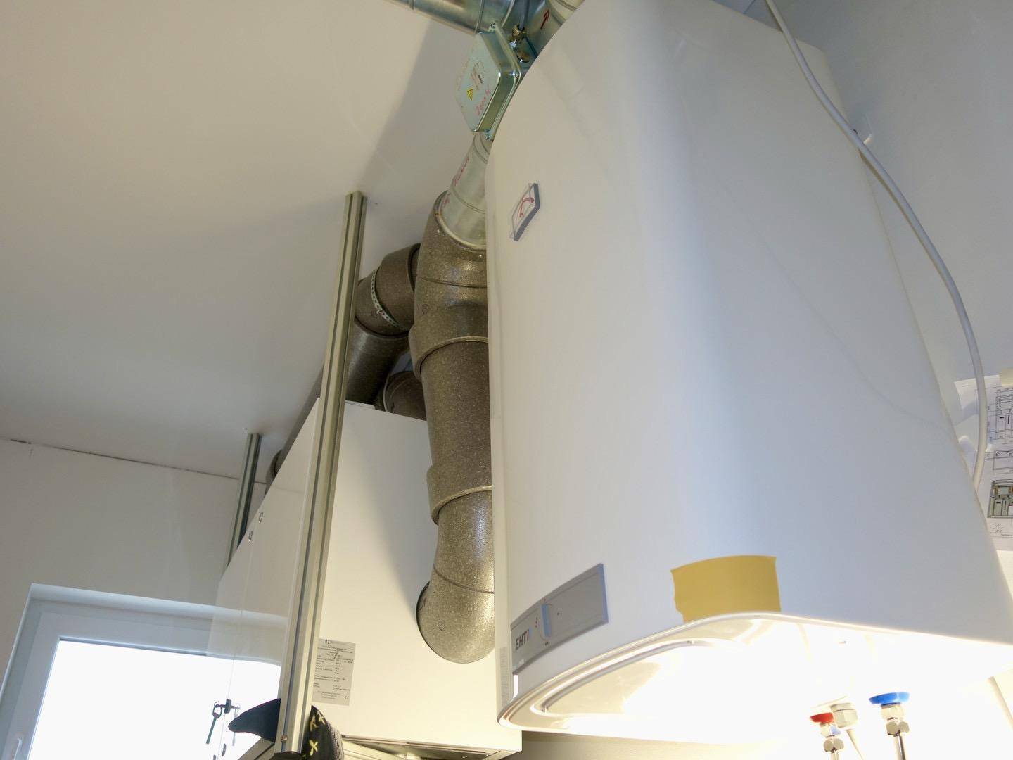 Technischer Raum mit Warmwasserbereiter und sichtbaren Lüftungsrohren an der Decke.