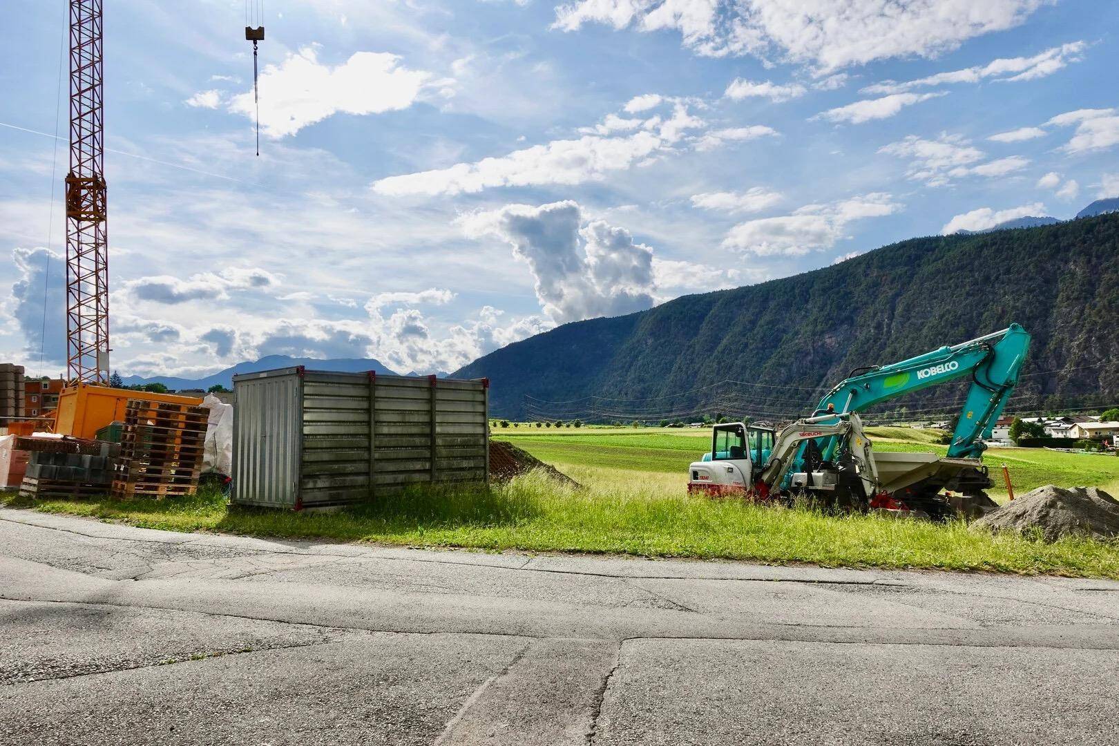 Baustelle mit Bagger, Kran und Containern vor einer weiten Landschaft mit Bergen.