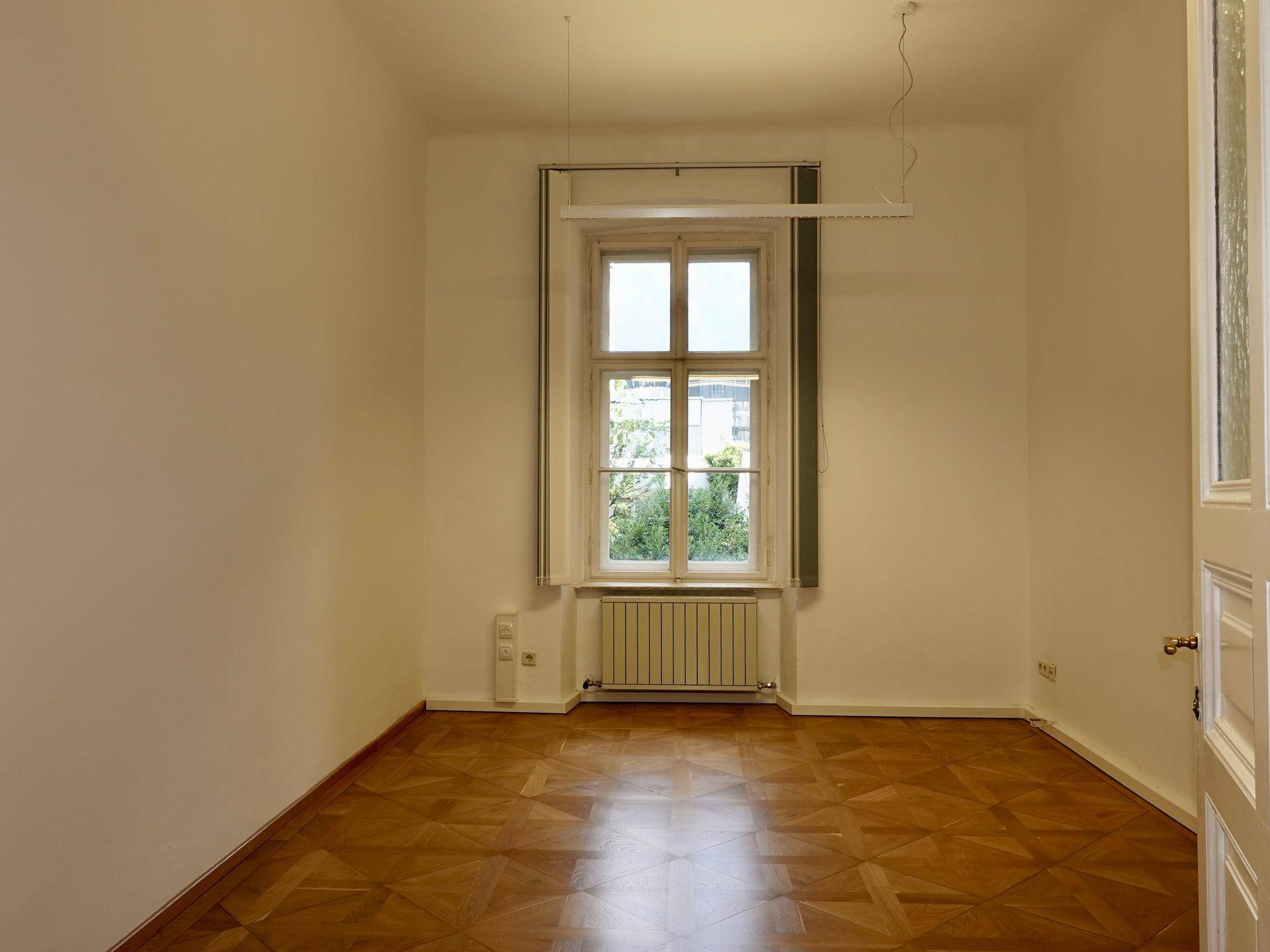 Leerer Raum mit Fischgrätparkett, einem großen Fenster und einem Heizkörper darunter, sehr hell.