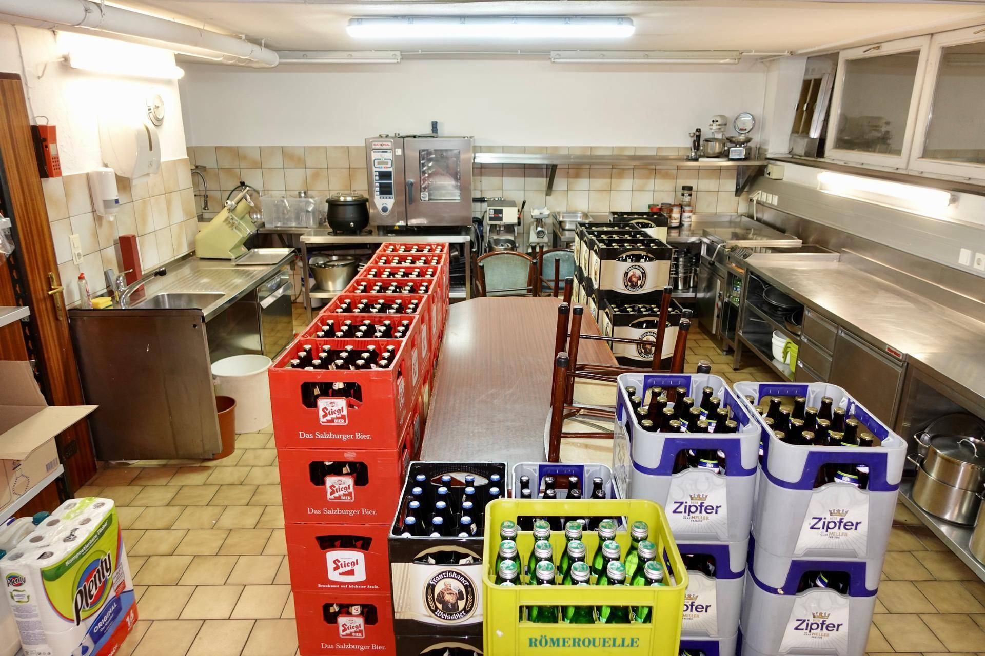Geräumiger Lagerbereich mit zahlreichen Getränkekisten, einem Arbeitstisch und professionellen Küchengeräten im Hintergrund.