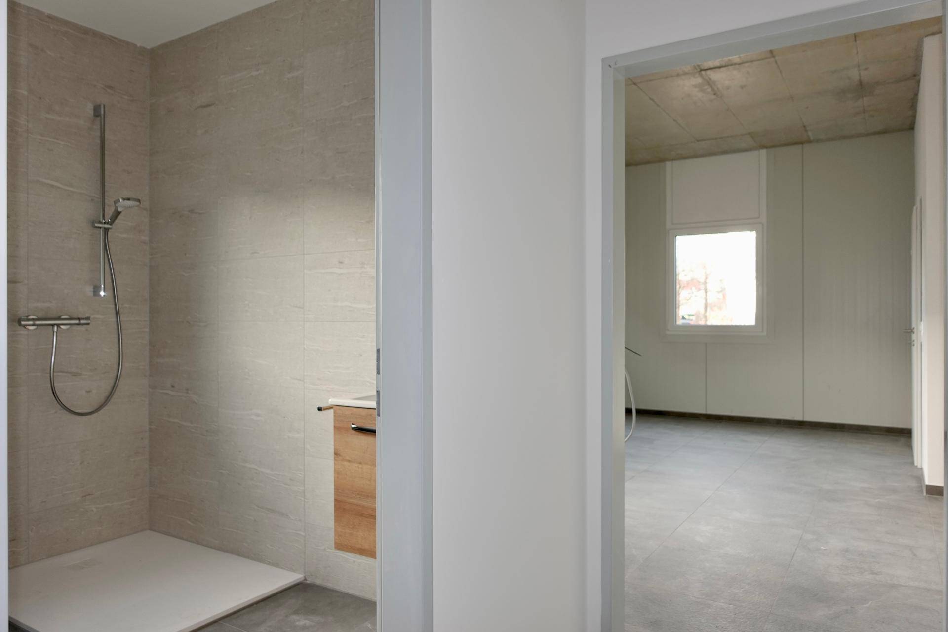 Modern geflieste Duschkabine mit Armaturen und Blick in einen angrenzenden Raum mit Fenster.