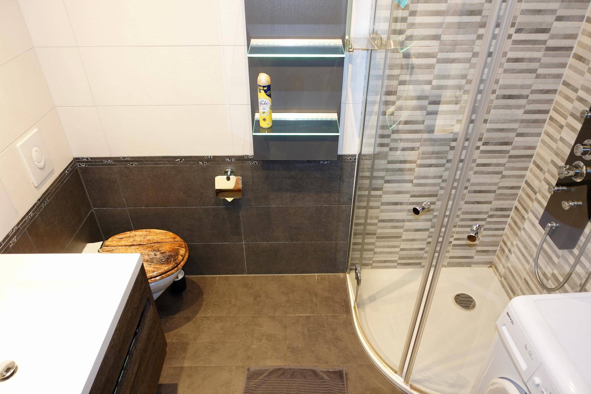 Modernes Badezimmer mit Duschkabine, dunklen Fliesen und integrierten Ablagen.