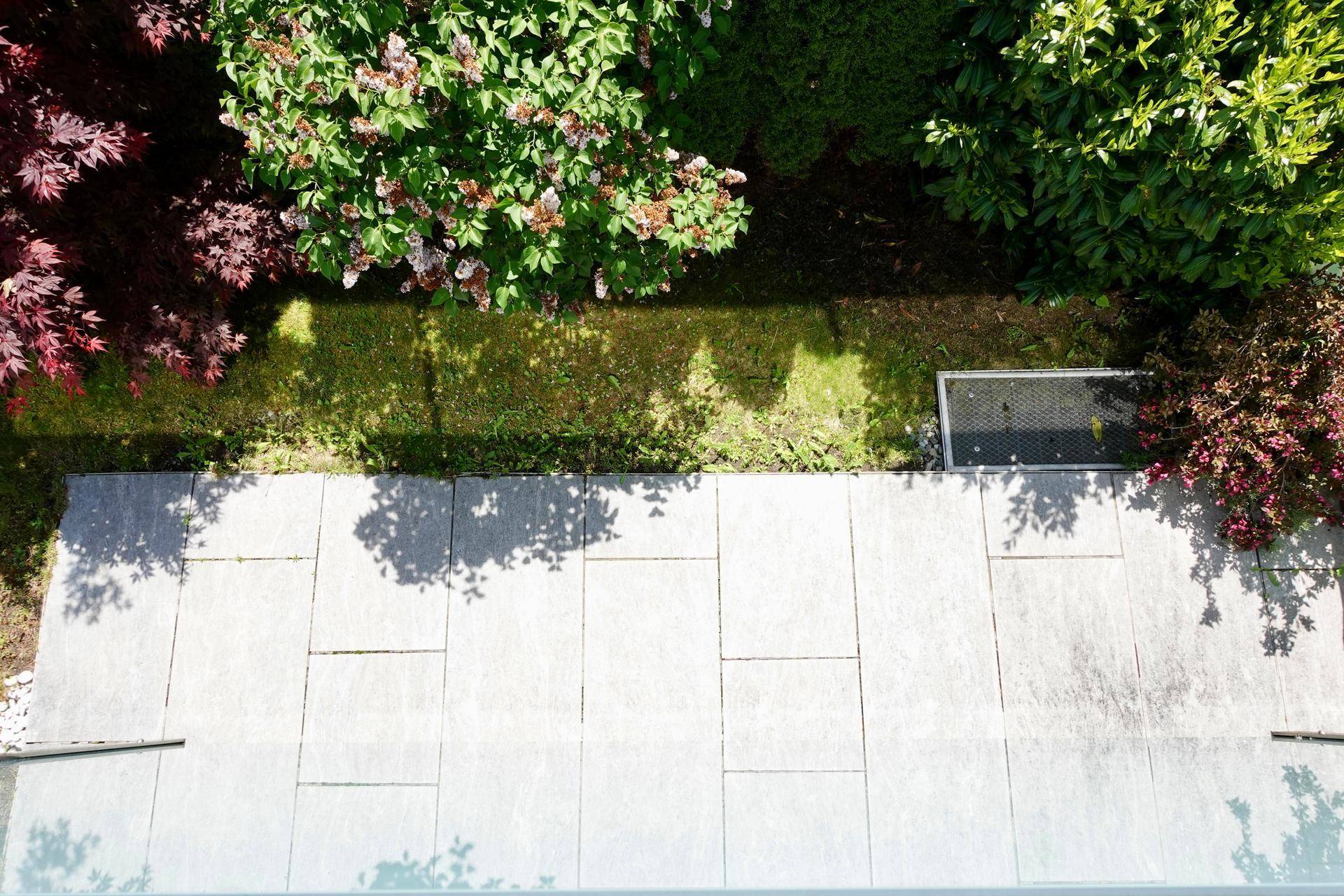 Gepflasterte Terrasse von oben gesehen, angrenzend an einen grünen Gartenstreifen mit Sträuchern.