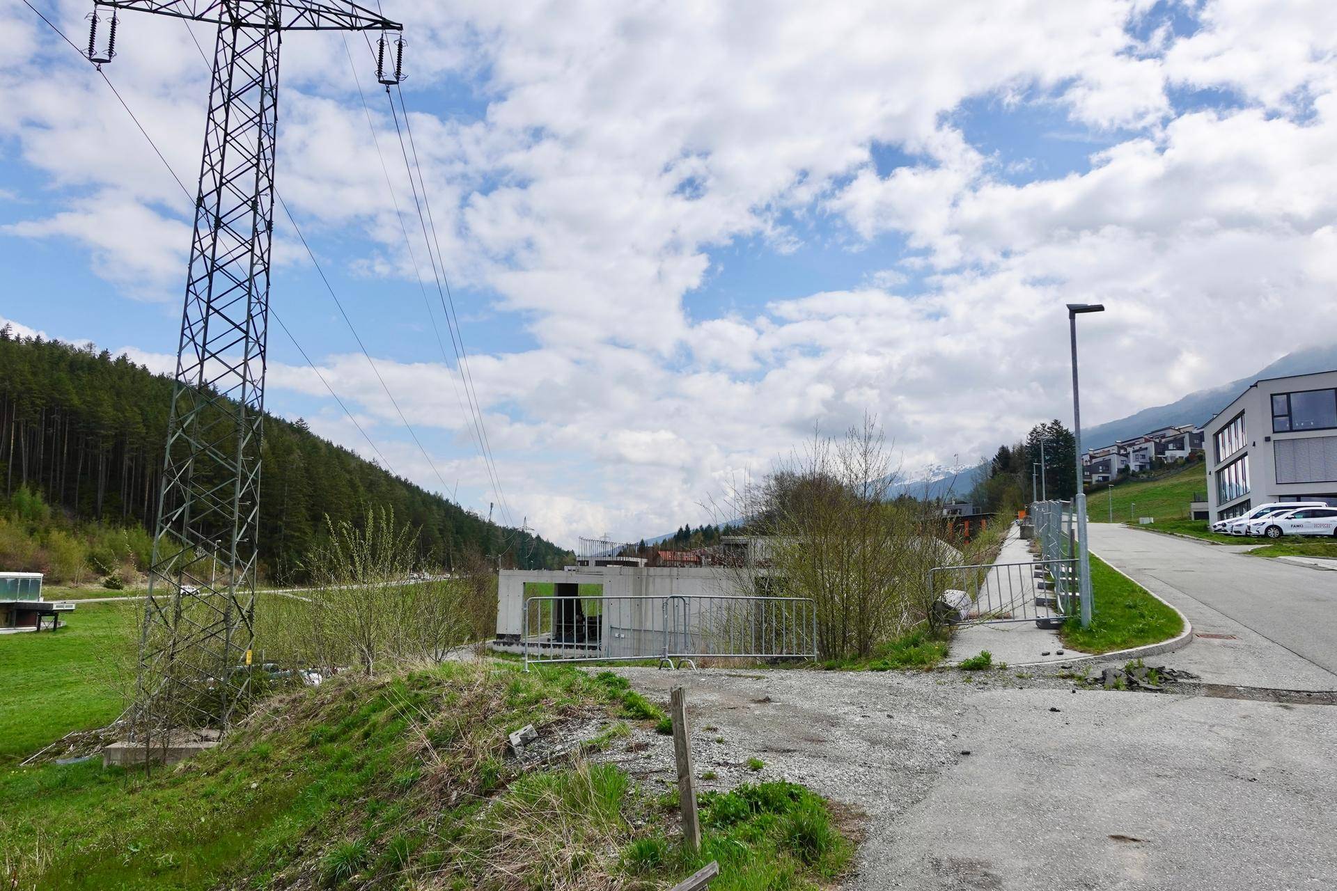 Straße führt an einem modernen Gebäude vorbei, mit einem großen Strommast im Vordergrund und grünen Hügeln und Bergen im Hintergrund.