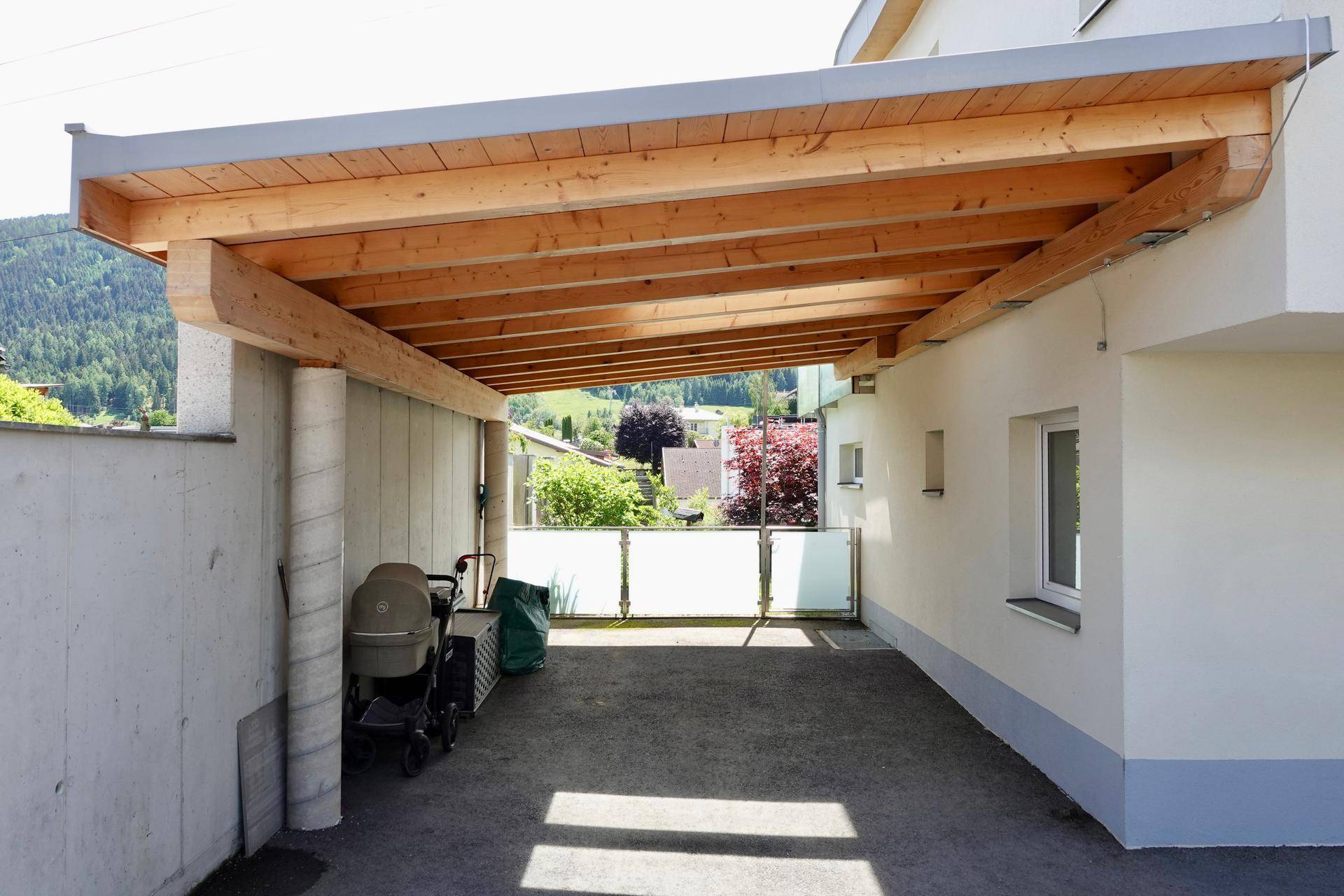 Geräumiger Holz-Carport mit Betonstützen und Blick auf die grüne Umgebung und Berge.