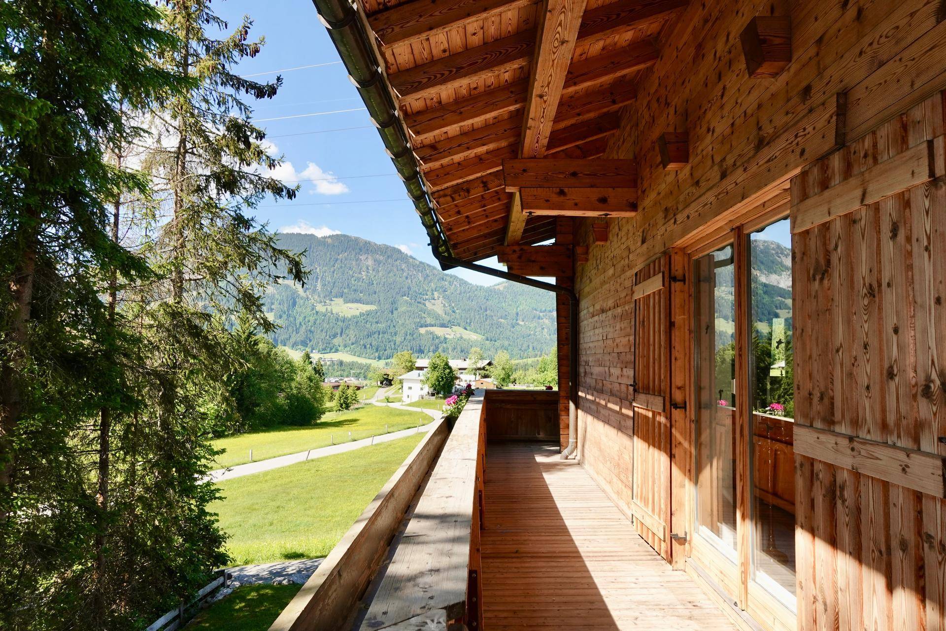 Langer Holzbalkon mit Blick auf die grüne Landschaft und die umliegenden Berge.