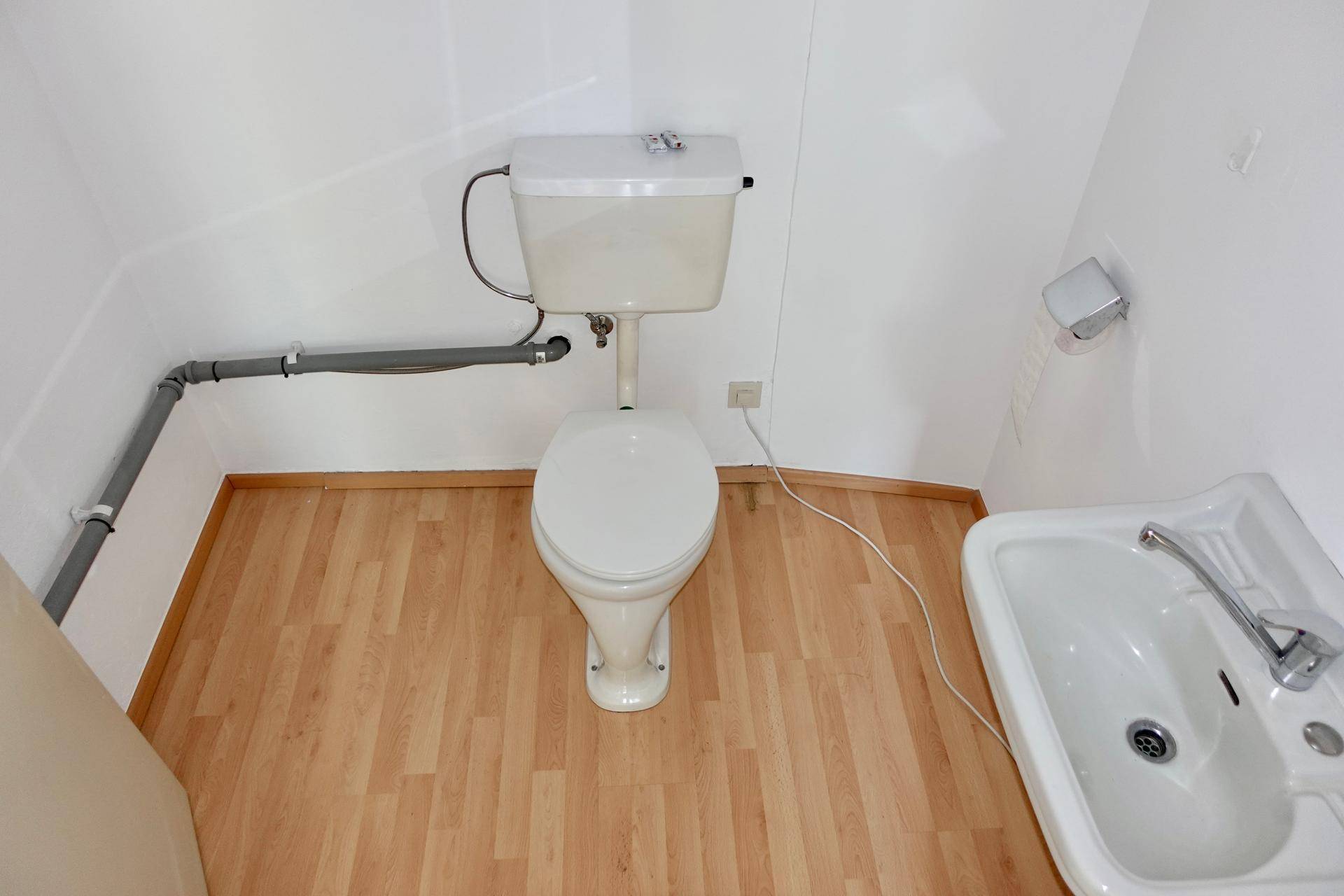 Kleine Gästetoilette mit WC, Waschbecken und sichtbaren Rohrleitungen an der Wand.