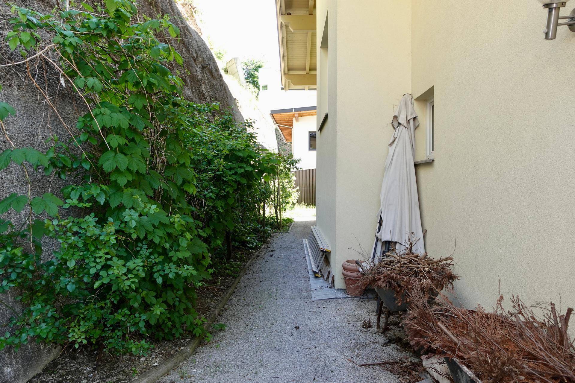 Schmaler Weg neben dem Haus mit üppigem Grün und Gerümpel.